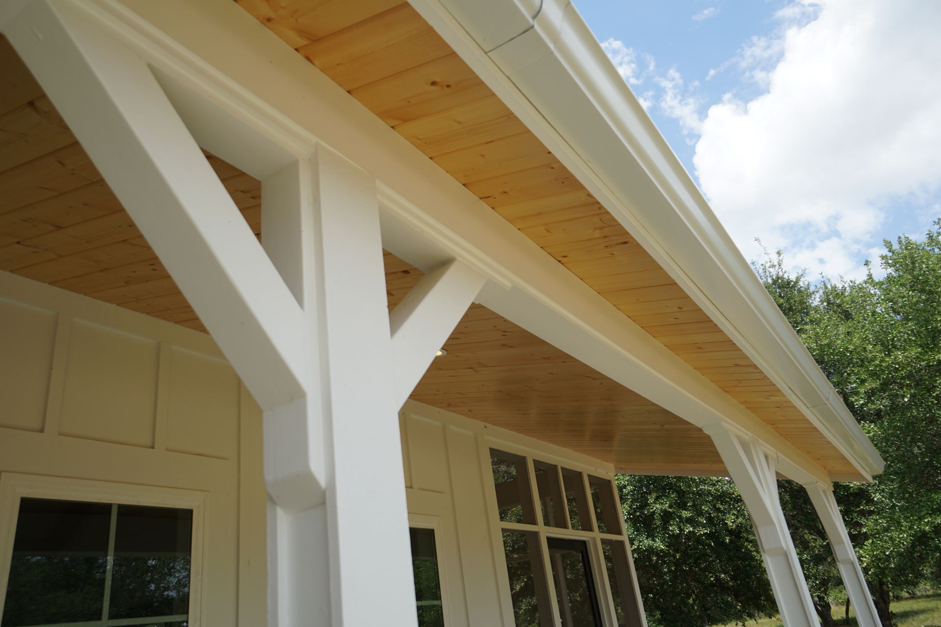 White porch posts and beams supporting a wooden ceiling and gutter; house exterior.