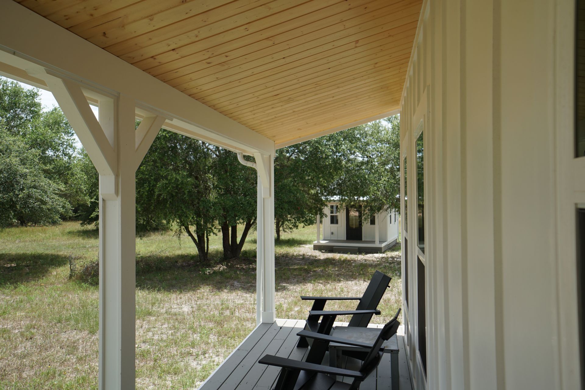 Porch with white columns, wood ceiling, two chairs, and a small building in the background.