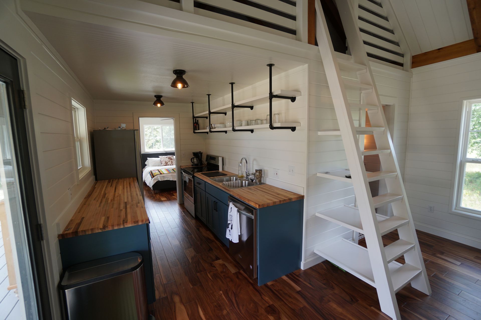 Interior view of a tiny home with a kitchen, loft ladder, and bed. Wooden floors, white walls, and blue cabinets.