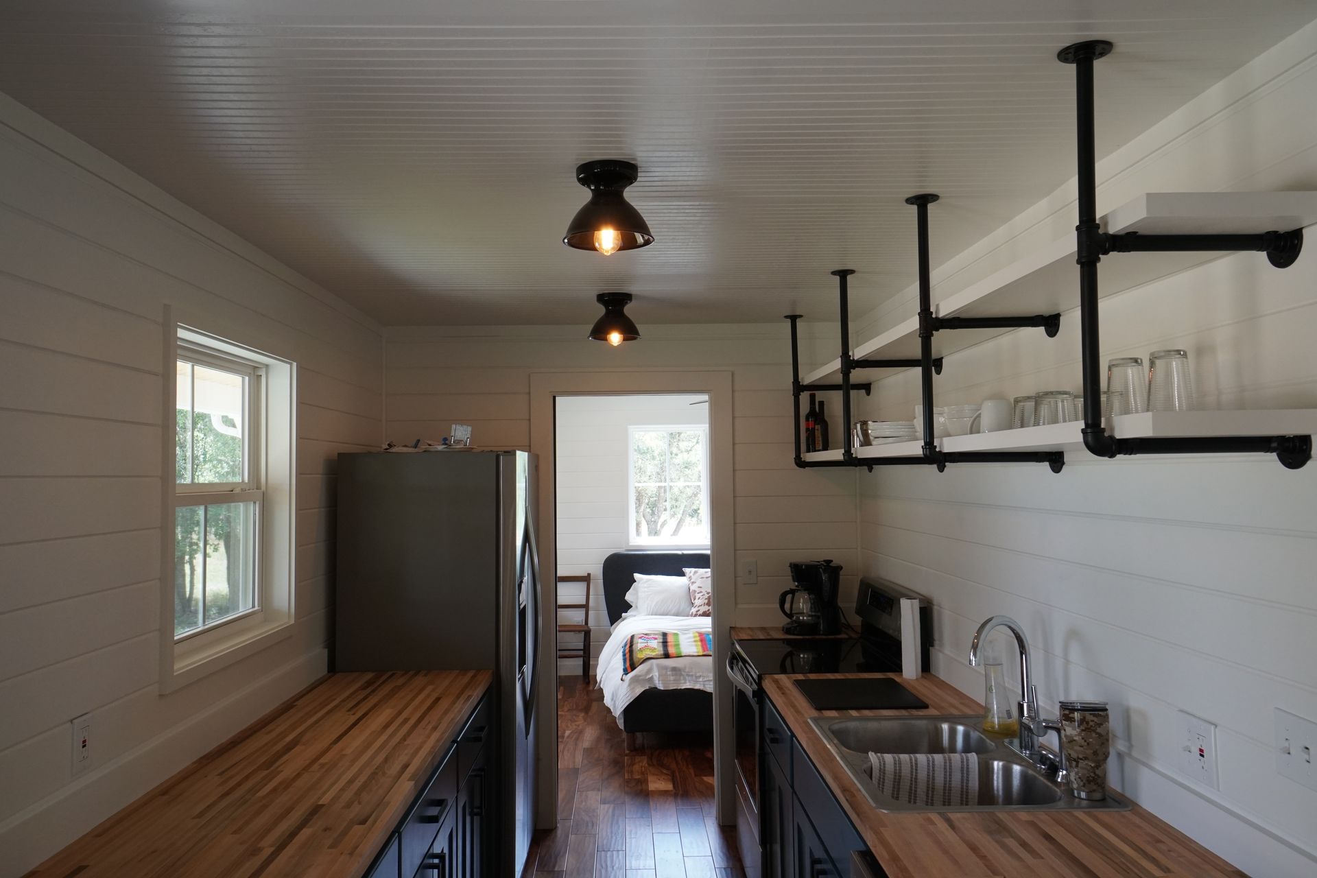 Interior view of a tiny home with a kitchen and bedroom. White walls, wood countertops, and black pipe shelving.