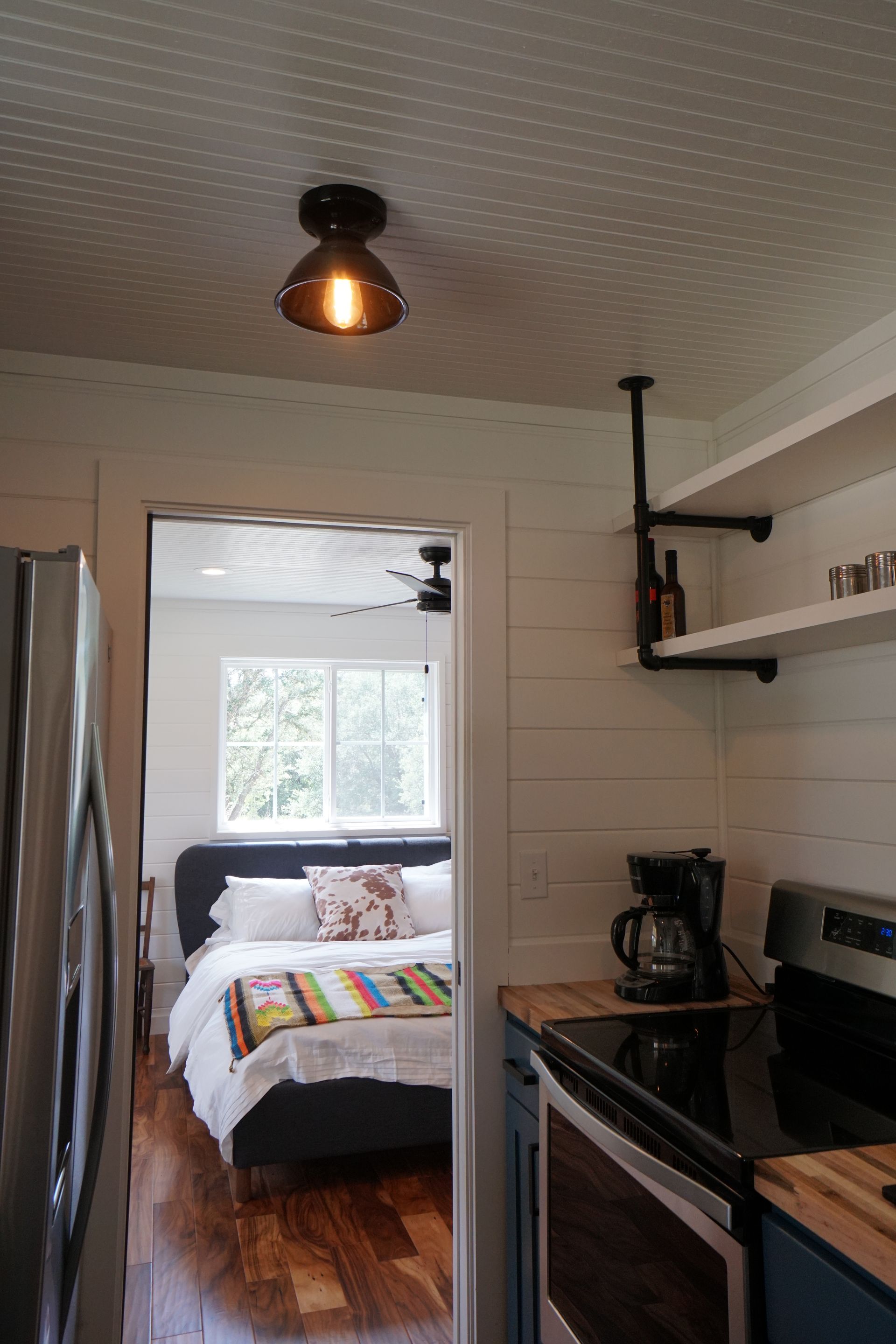 Small kitchen, bedroom view. White walls, wood floor. Bed with colorful blanket. Black stove and coffee maker.