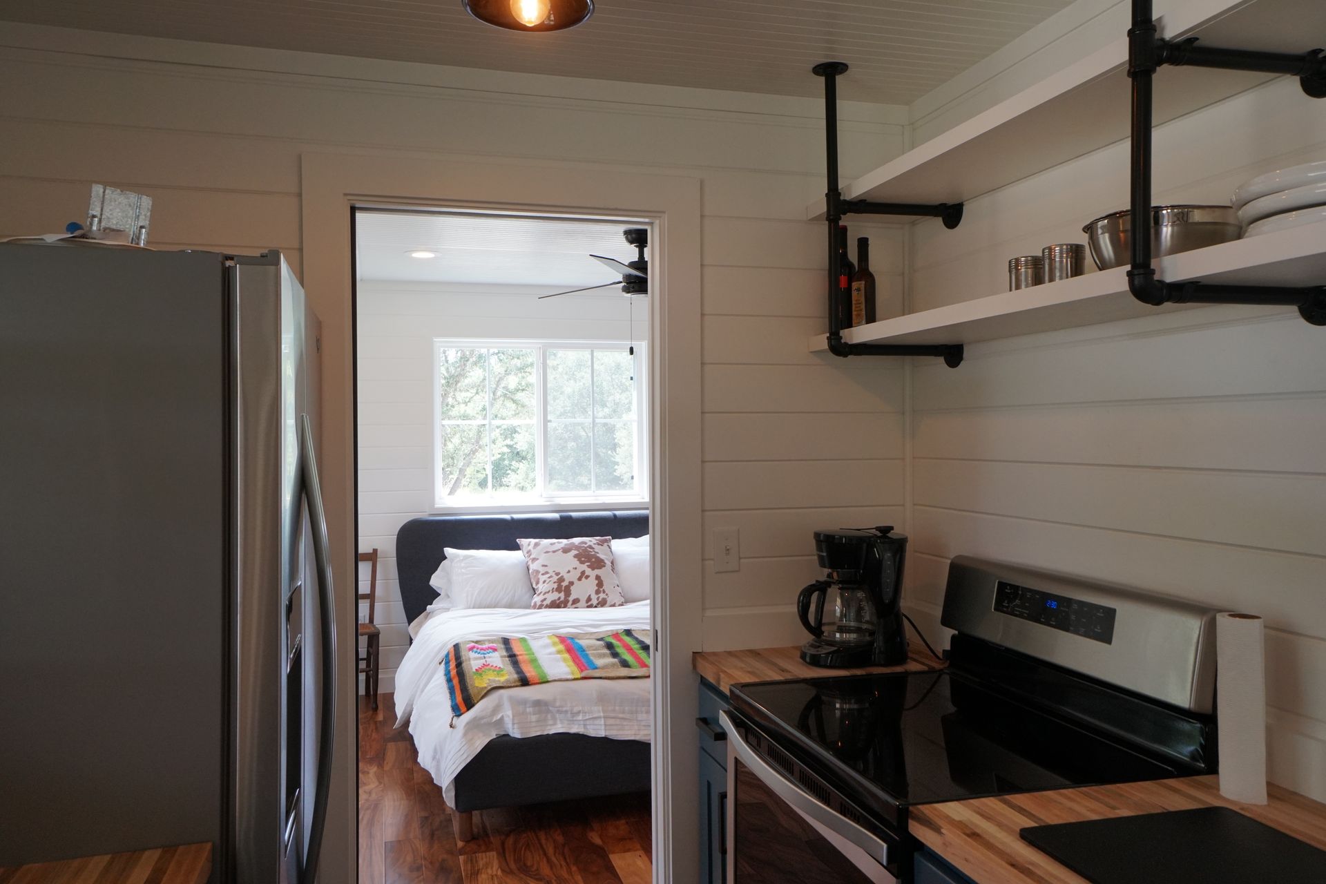 Kitchen with open shelves, stove, and a view into a bedroom with a bed and a window.