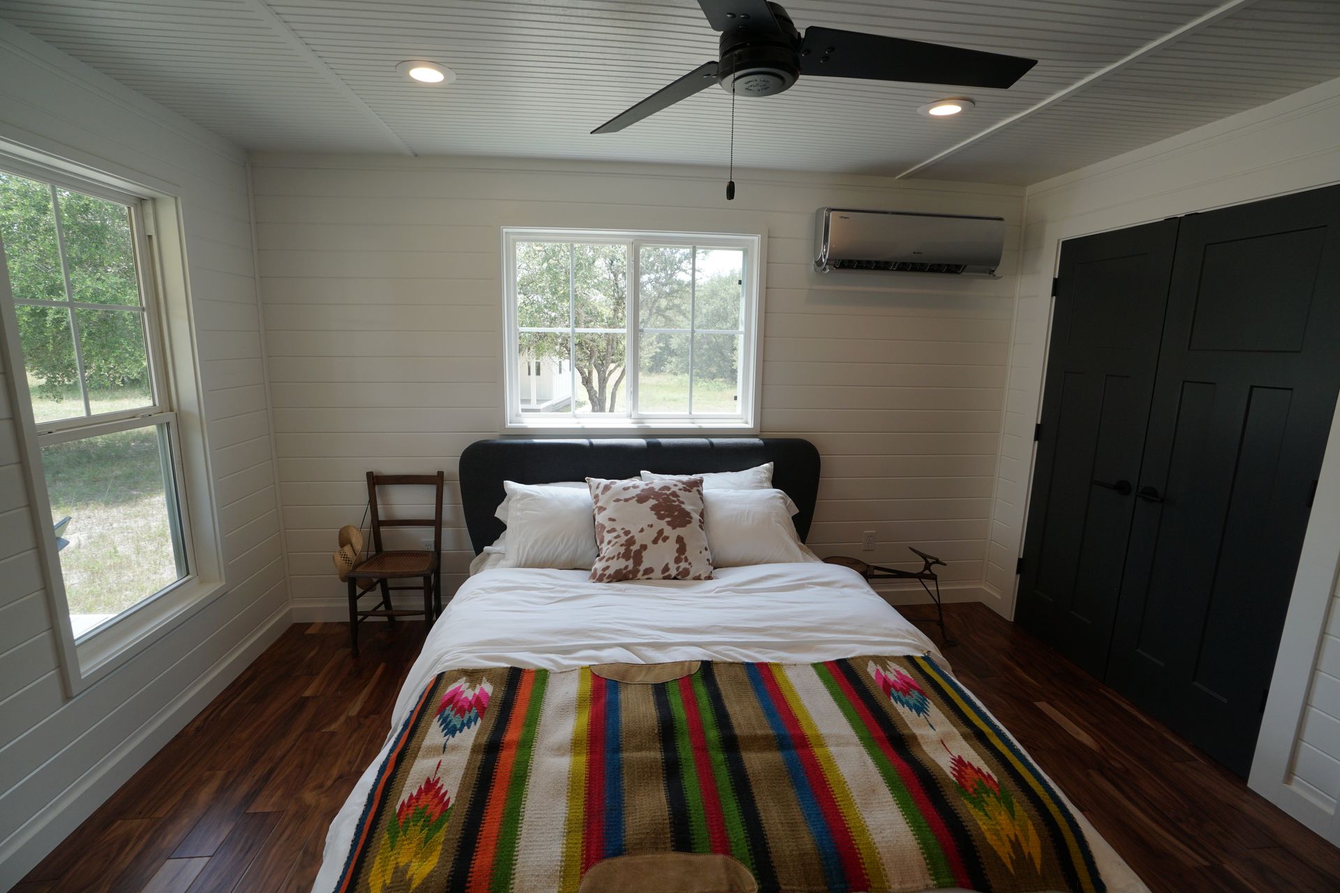 Bedroom with bed, closet, and windows. White walls, wood floor, colorful blanket, air conditioner.