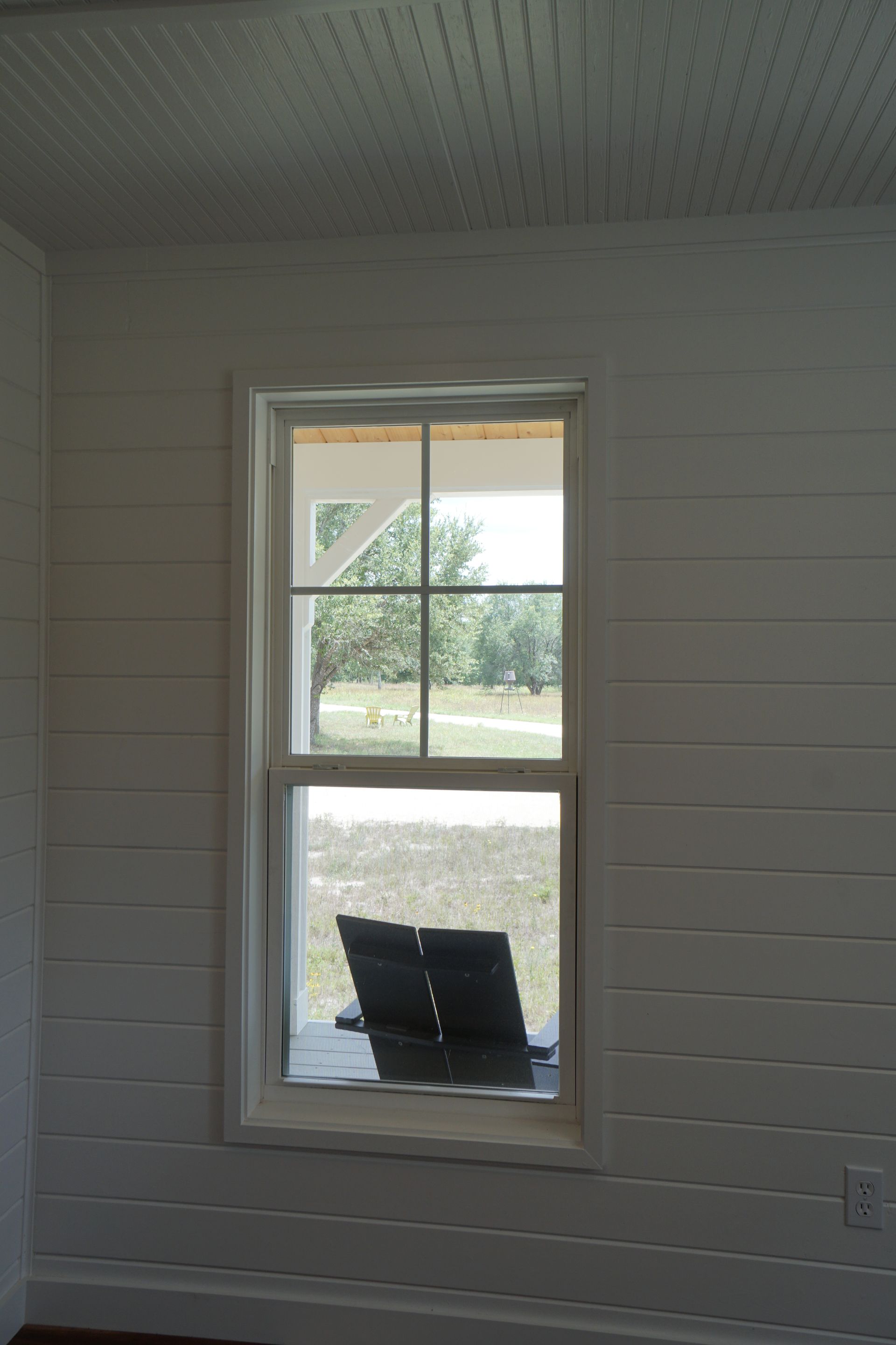 White-walled room with window. View of outside: trees, grass, and a dark object on a ledge.
