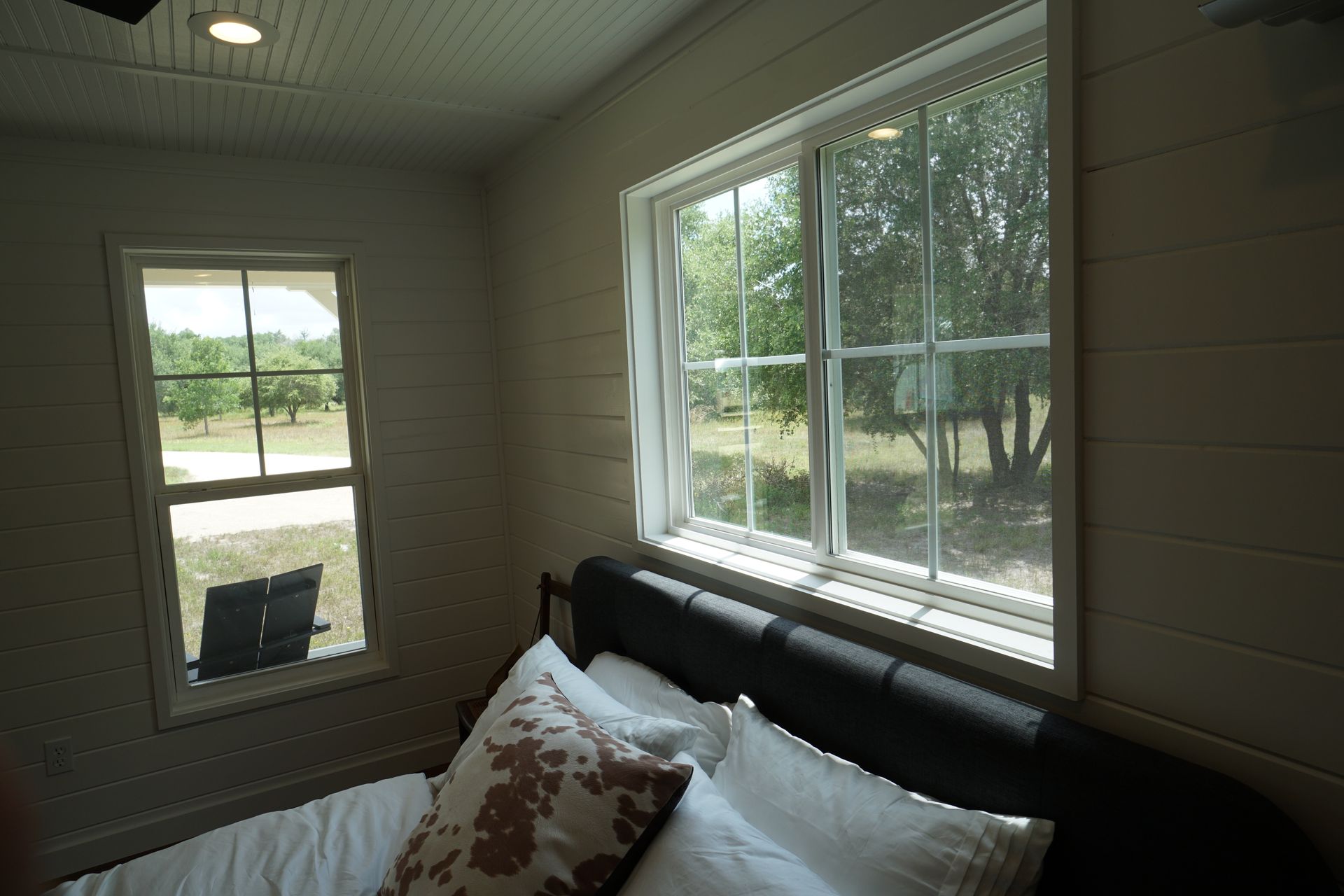 Bedroom with two windows overlooking a tree-filled outdoor space, with a bed and white walls.