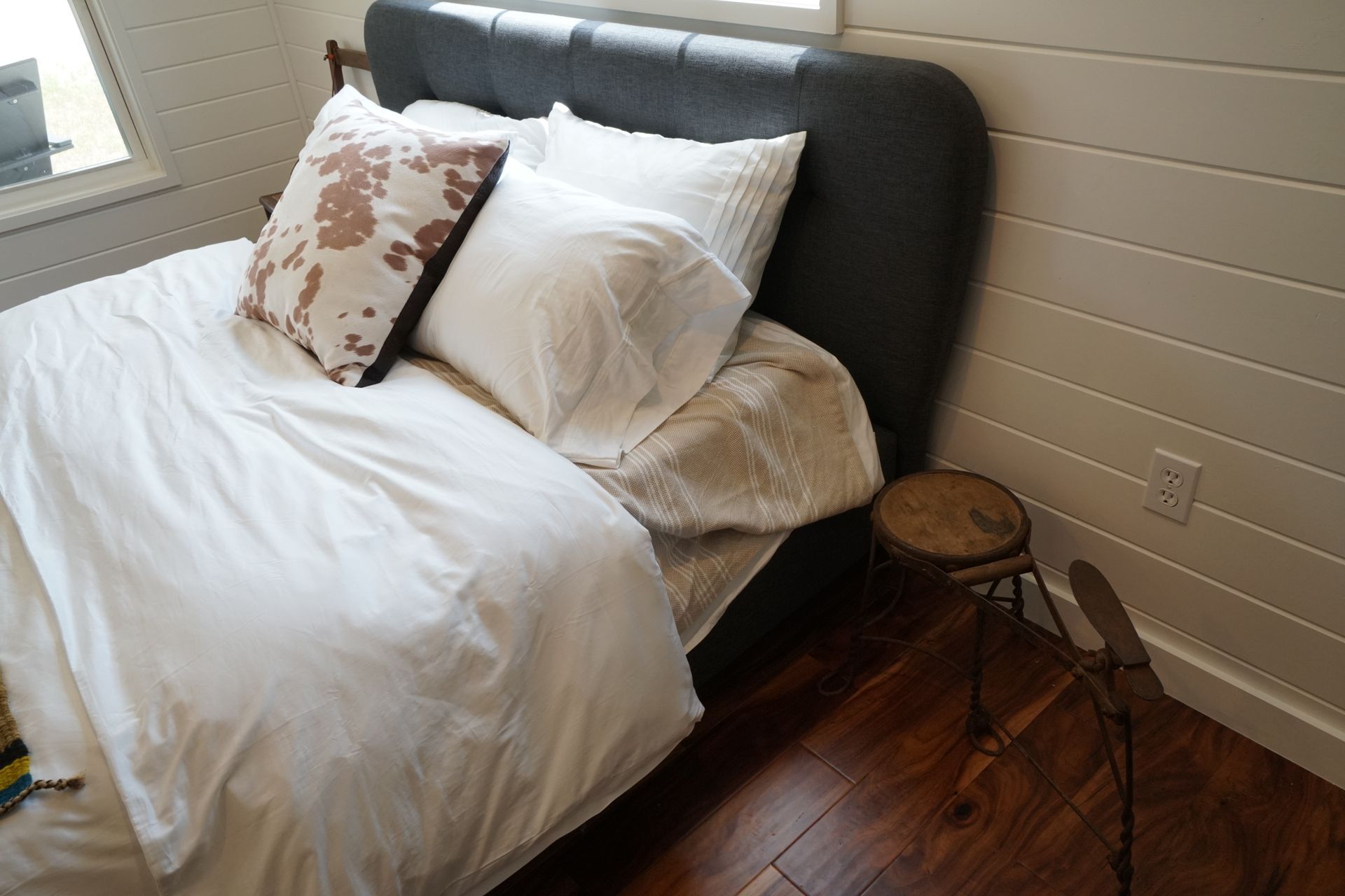 Bed with white linens, cowhide pillow, and a small stool on a hardwood floor.