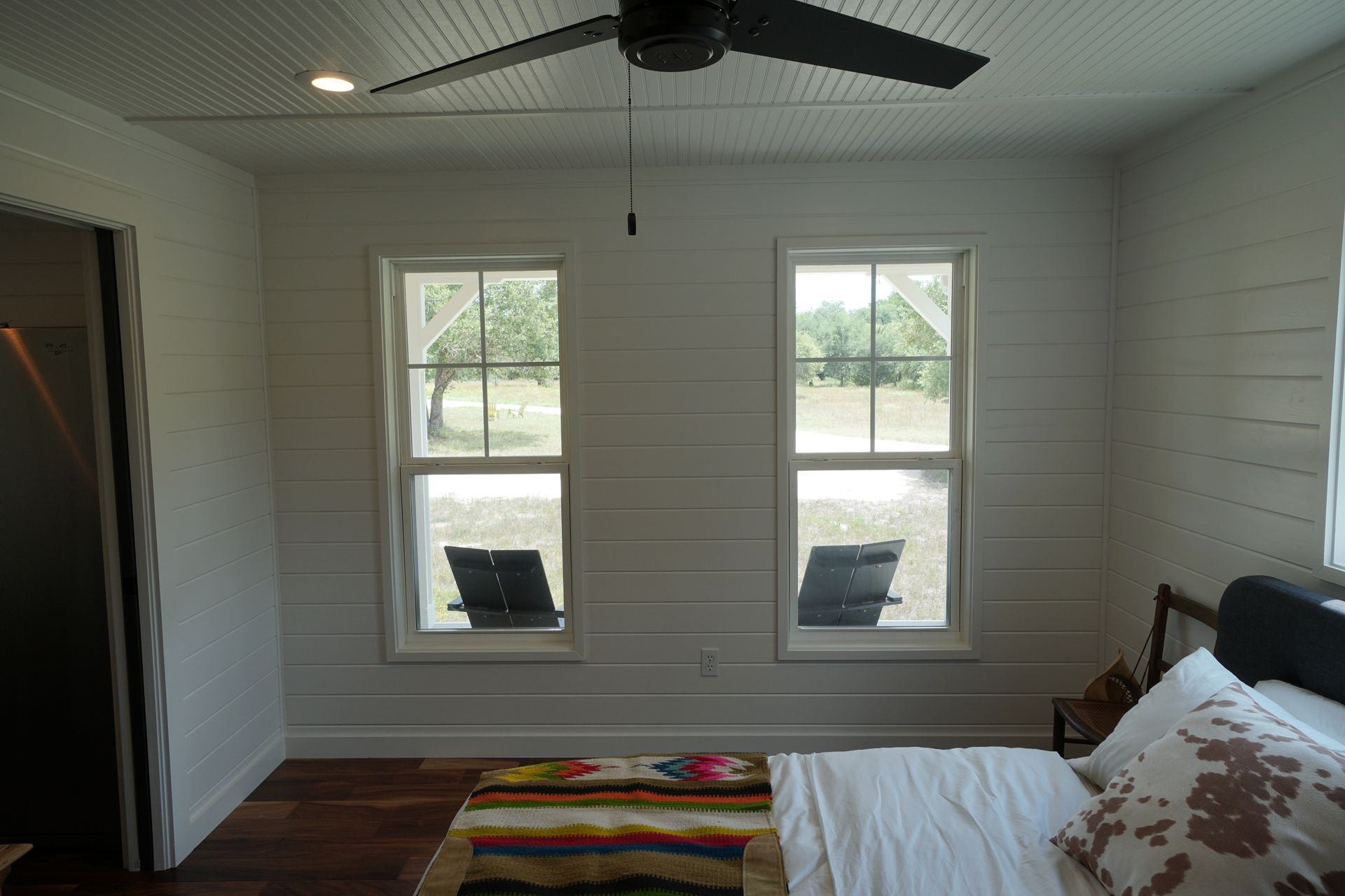 Bedroom with white horizontal wood paneling, two windows, ceiling fan, dark wood floors, bed, and a colorful rug.