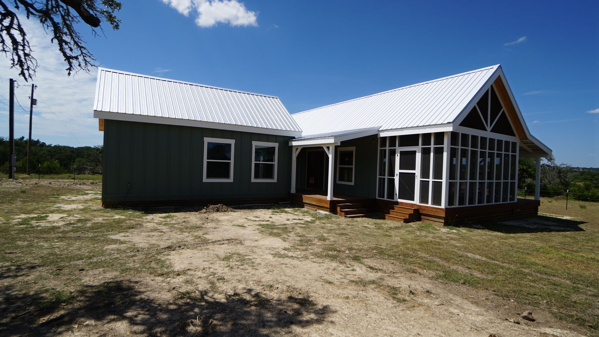 A house with a white metal roof and screened-in porch, painted green, sits on a dirt lot under a blue sky.
