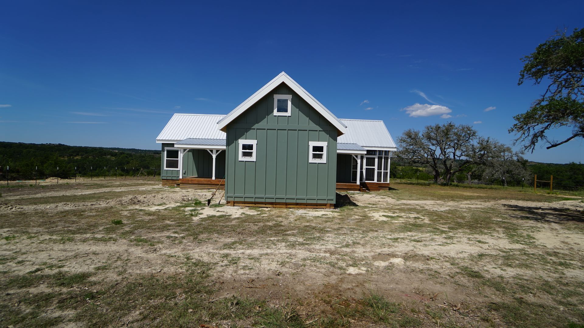 Green farmhouse with white trim and metal roof, on a dry, grassy plot under a blue sky.