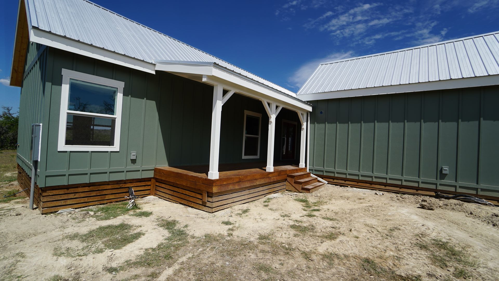 Green cabins with white trim, metal roofs, and wooden porch, set on a sandy lot under a blue sky.