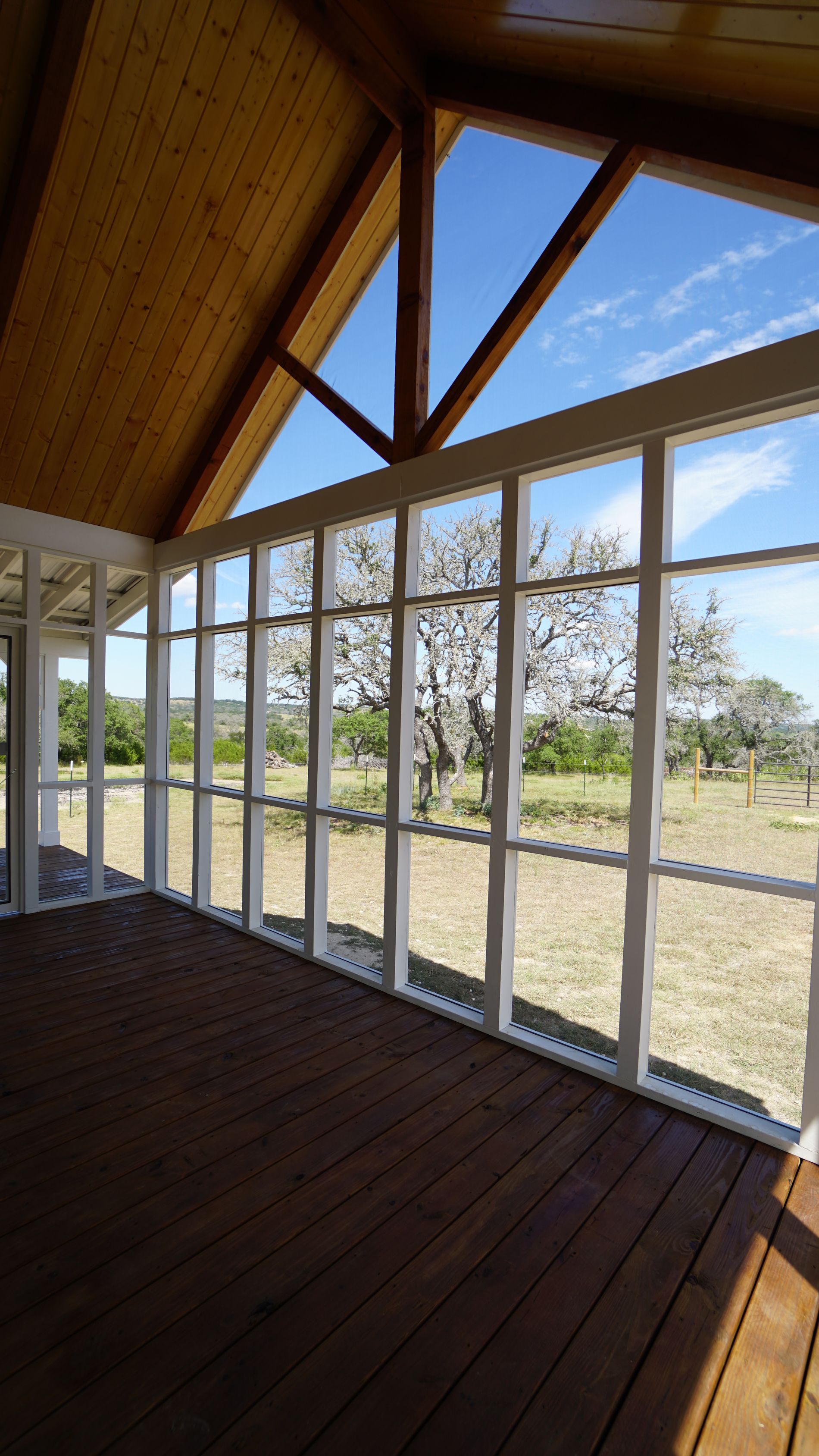 Sunroom with wood deck, framed windows, and a view of a tree and field under a blue sky.