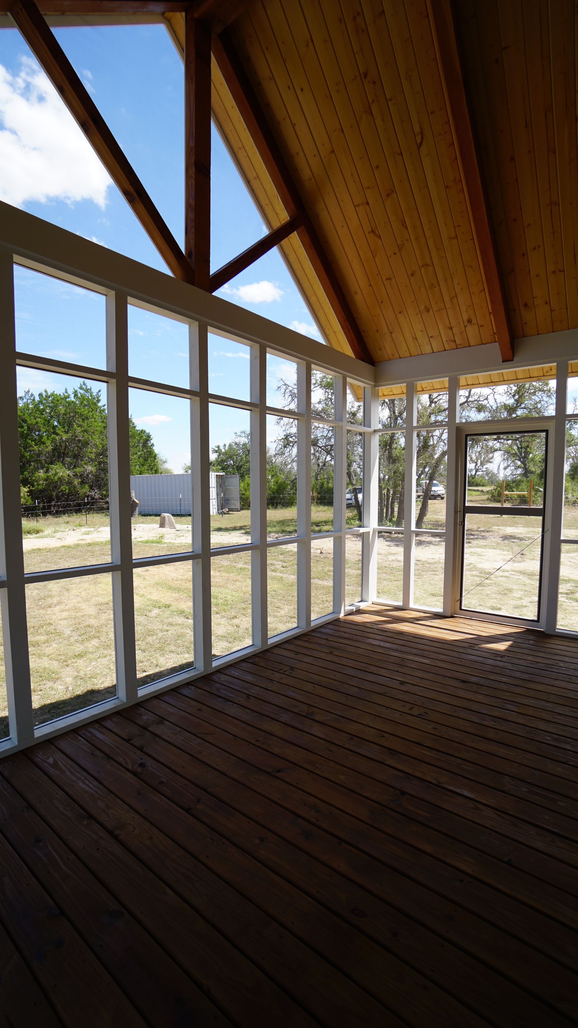 Wooden sunroom with large windows and a sloped, wood-paneled ceiling, overlooking an outdoor area.