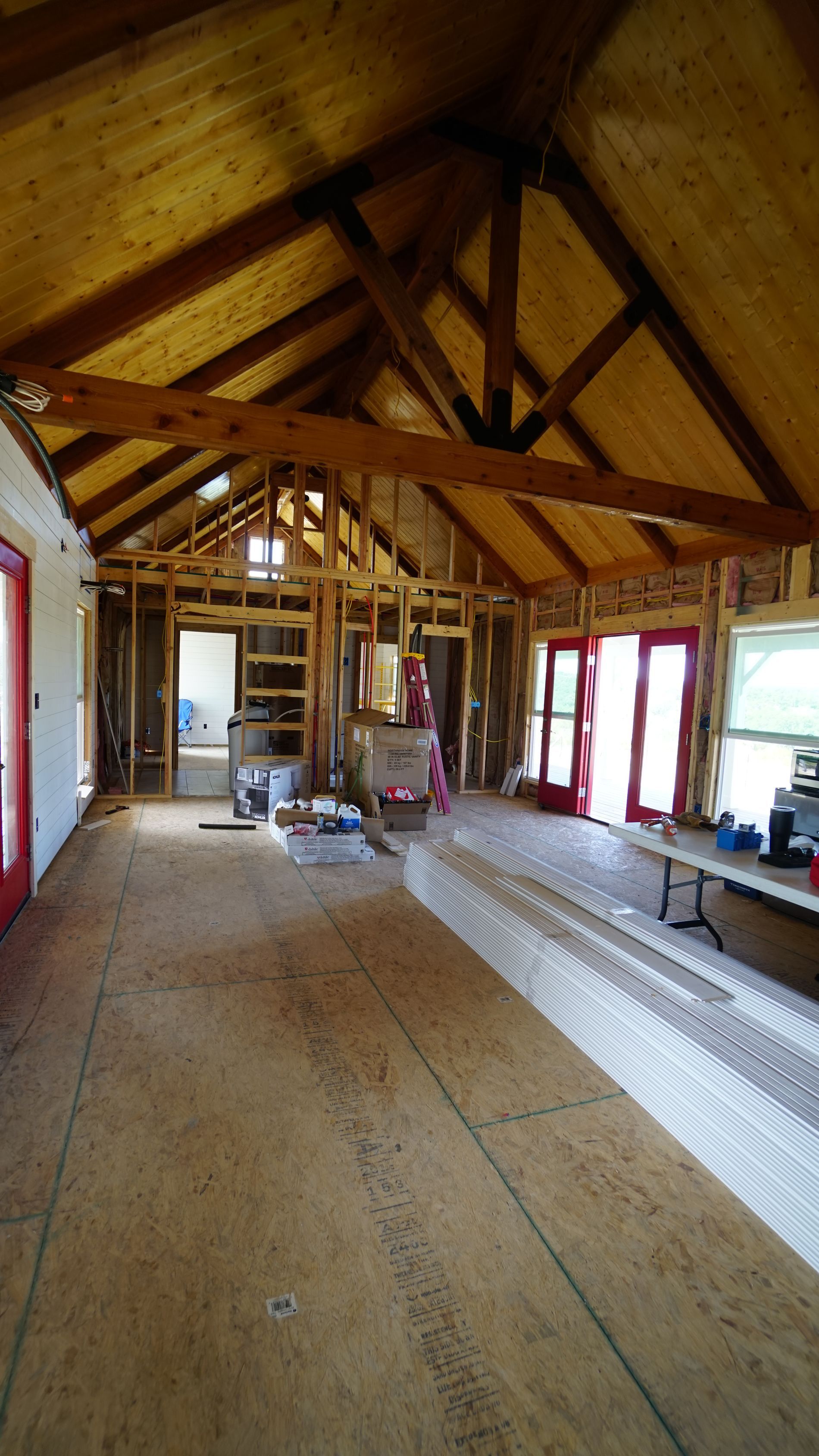 Interior construction site with exposed wooden framing and plywood floor. Red doors and windows visible.