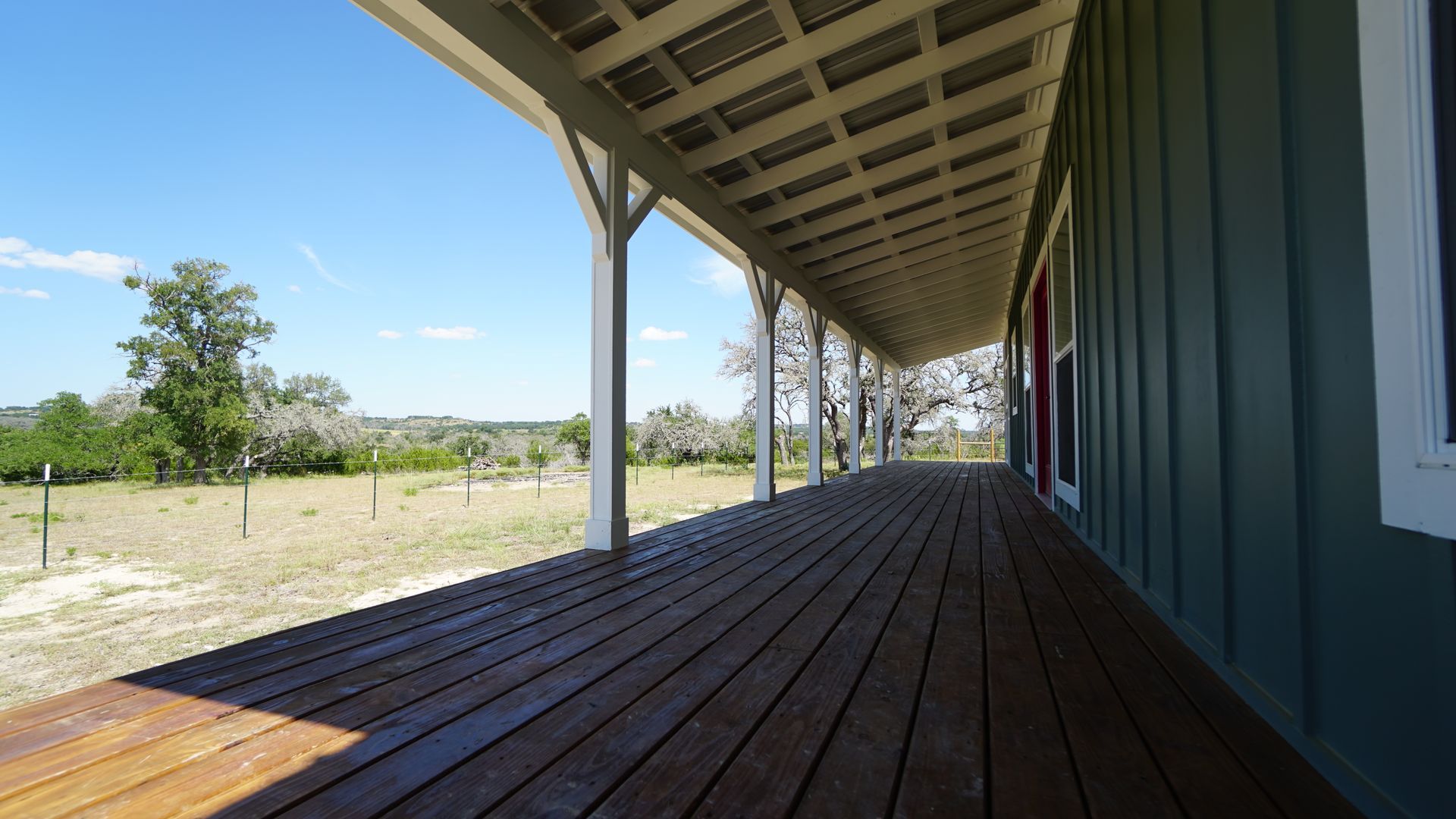 Wooden porch of a green house, overlooking a grassy field under a sunny sky.