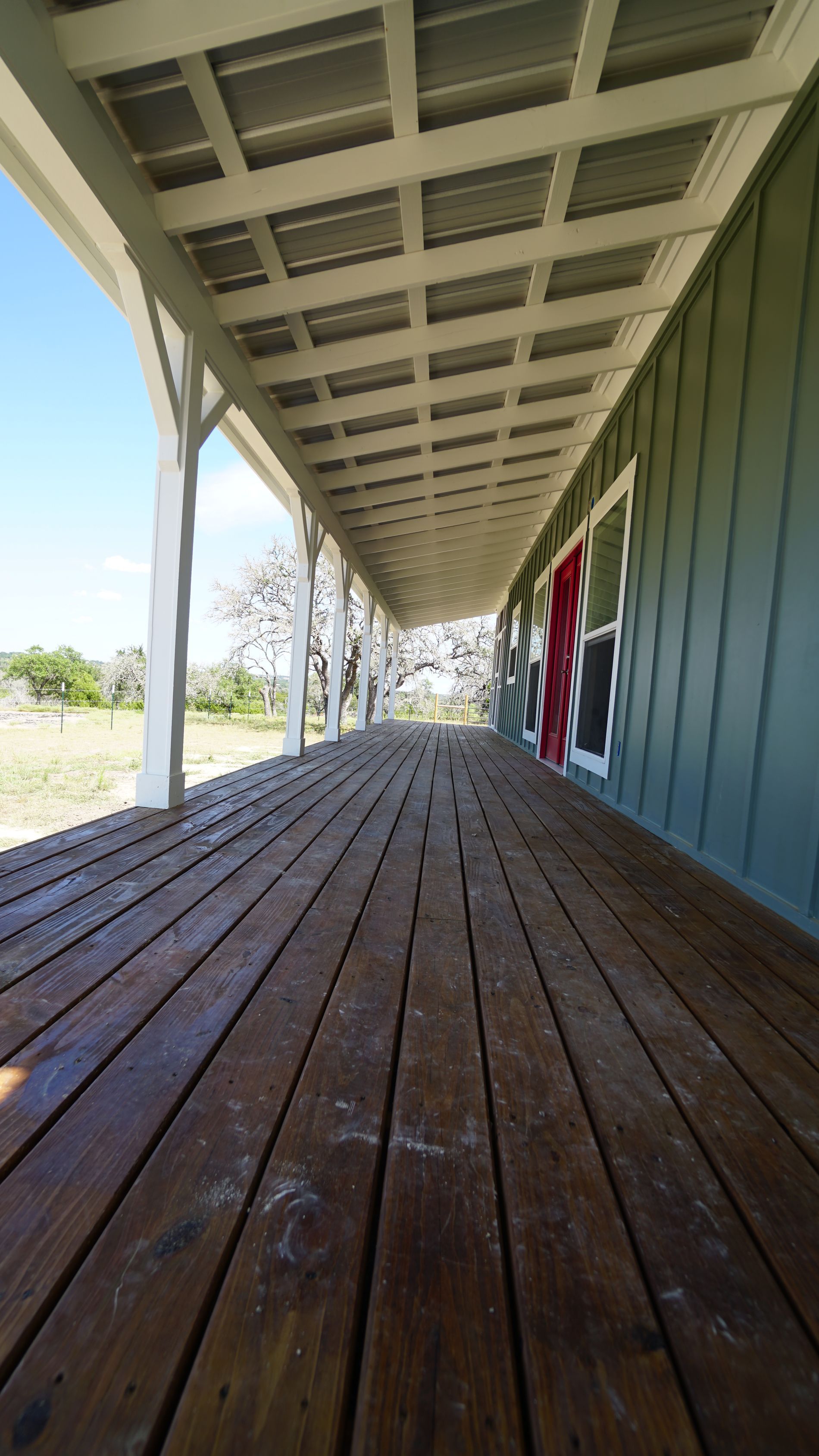 Wooden porch of a green house with a red door. White columns and ceiling, blue sky.