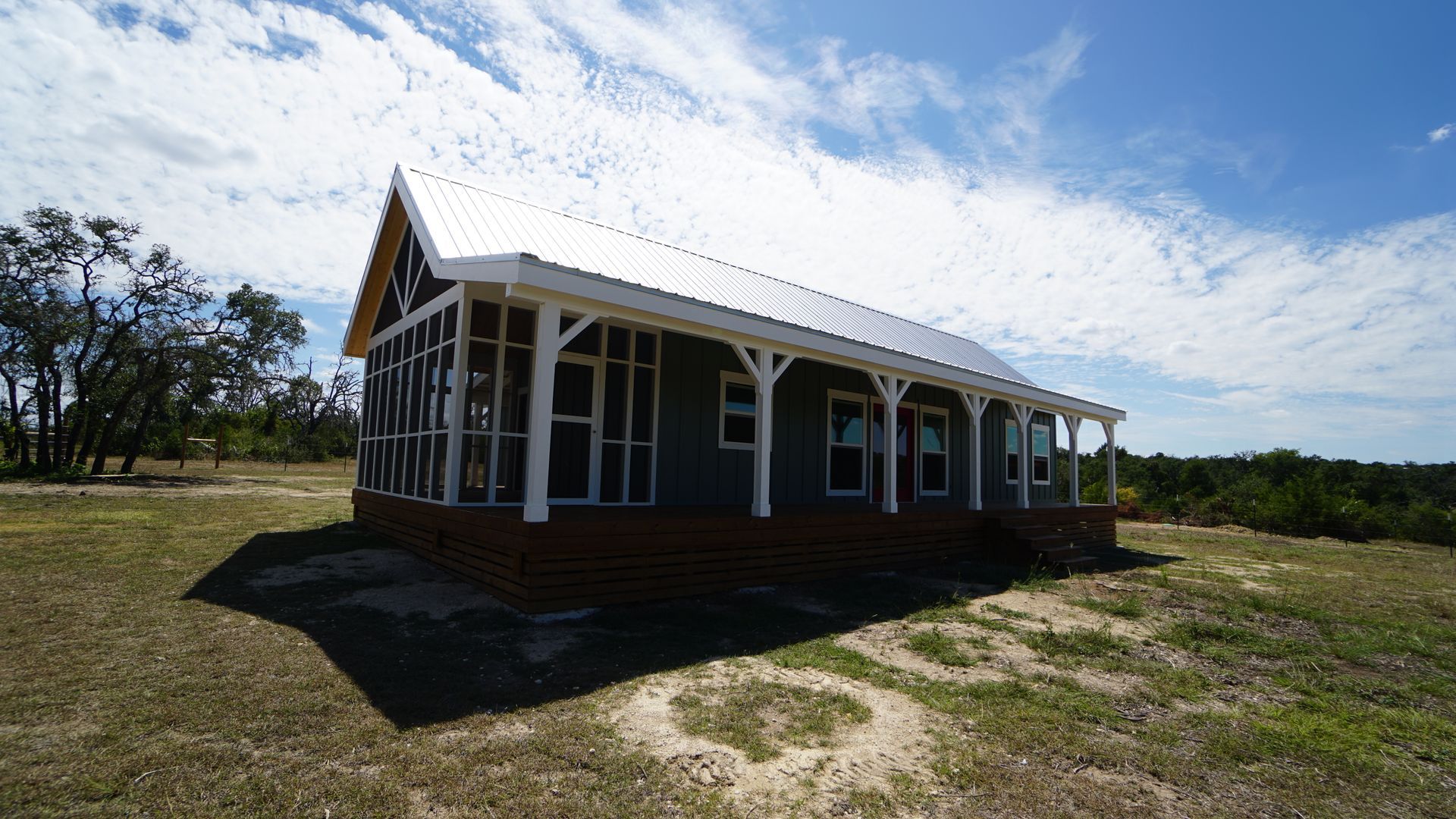 Small blue house with porch, white roof, and brick foundation, set in a grassy field under a sunny sky.