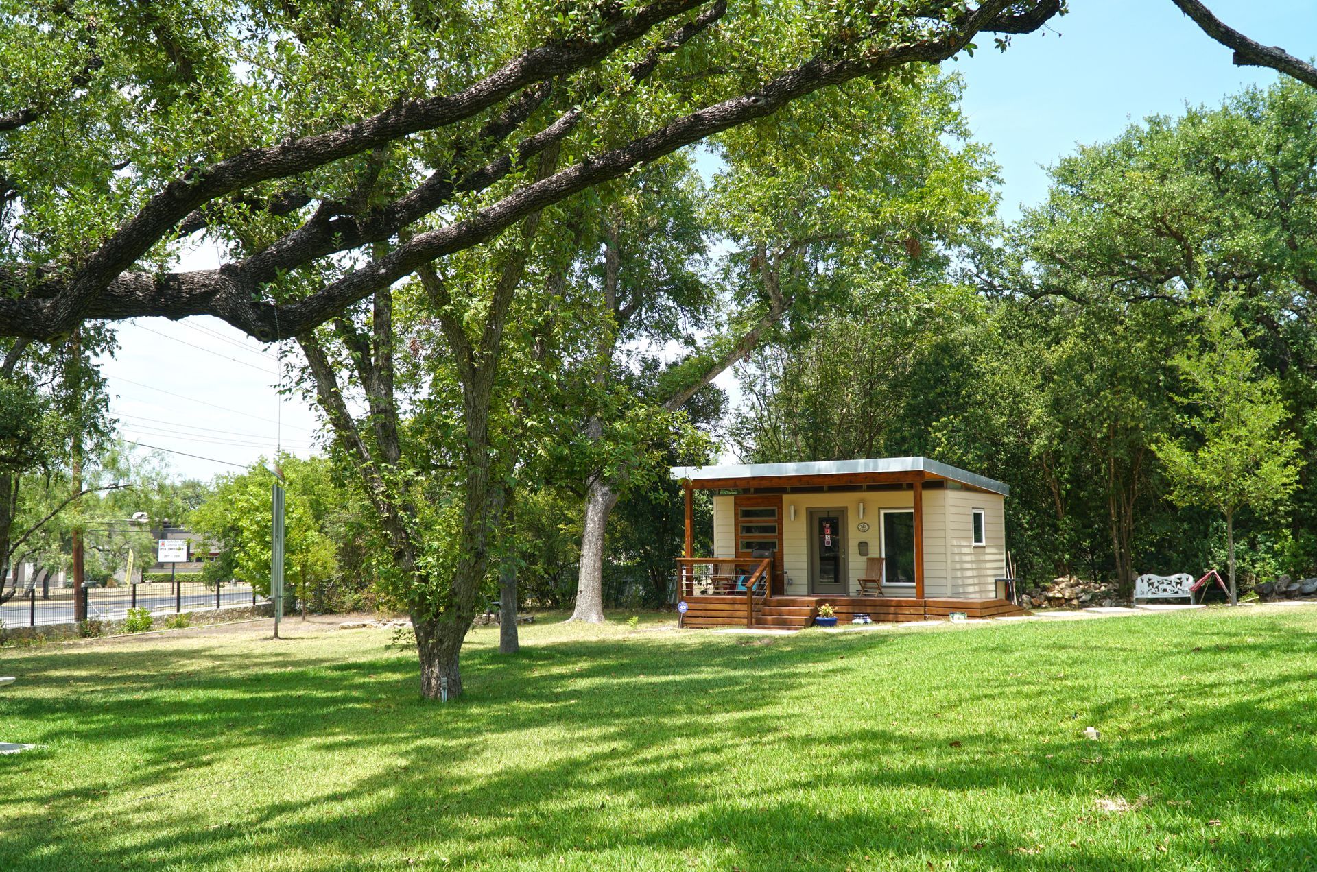Small cabin with porch in a grassy yard, under trees, near a road. Sunny day.