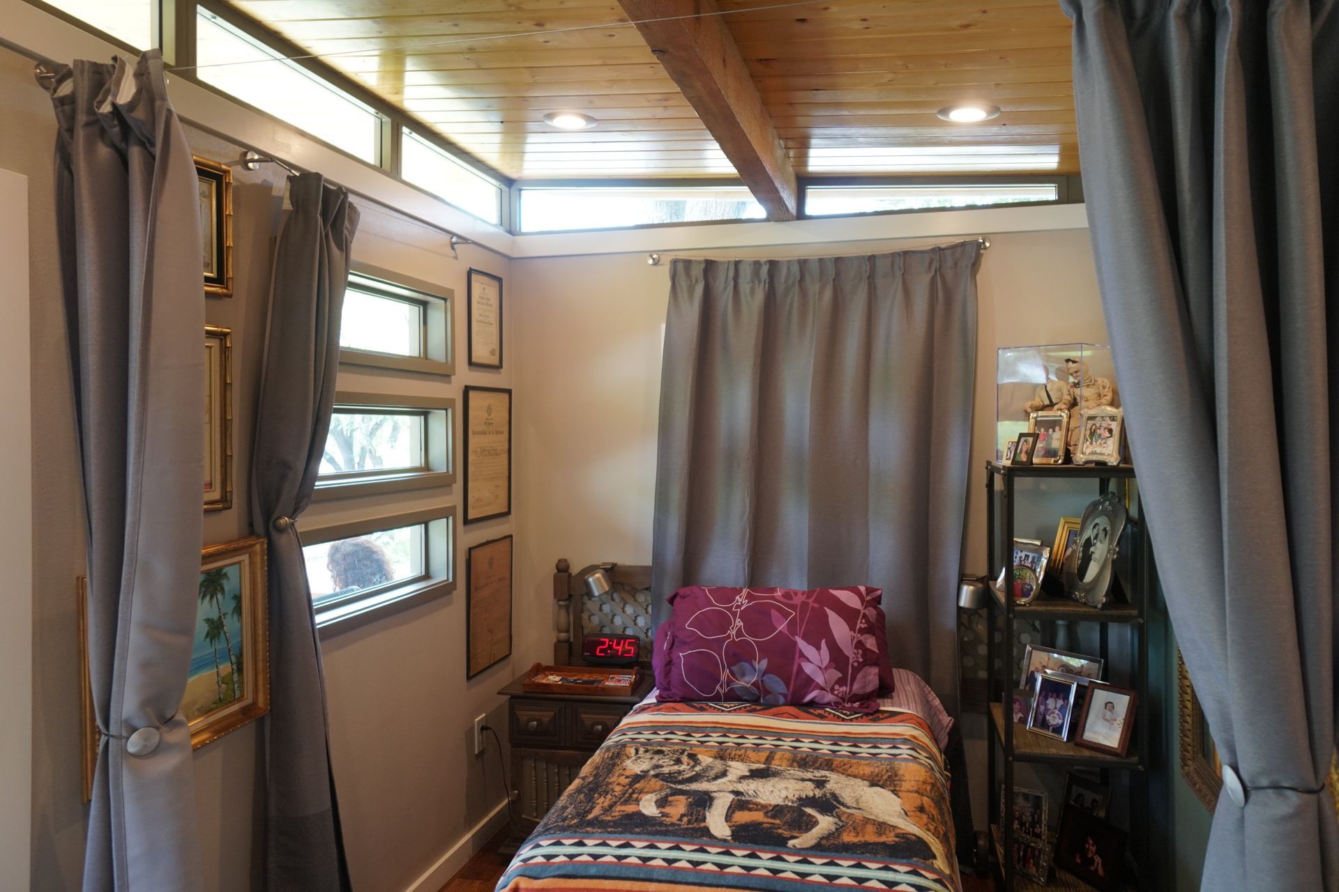 Small bedroom with bed, nightstand, shelf, windows, and gray curtains.