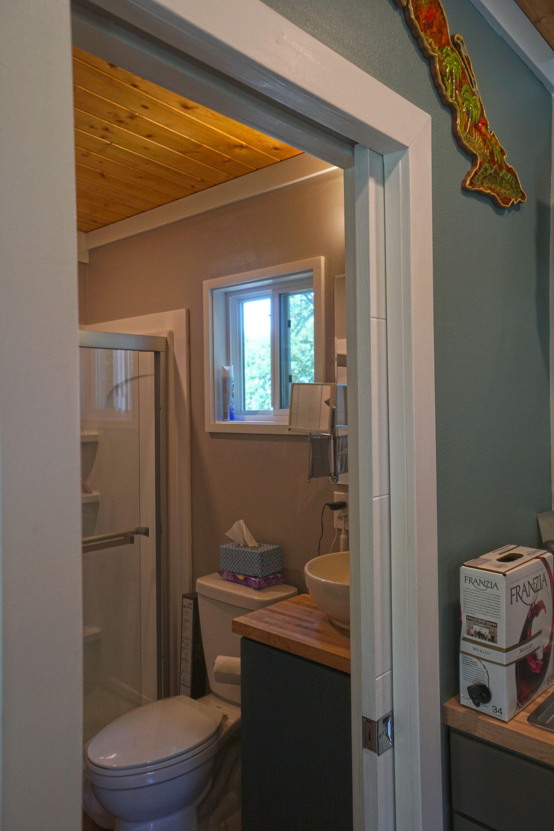 Bathroom doorway view: toilet, sink, shower, window with light blue wall, and wooden ceiling.