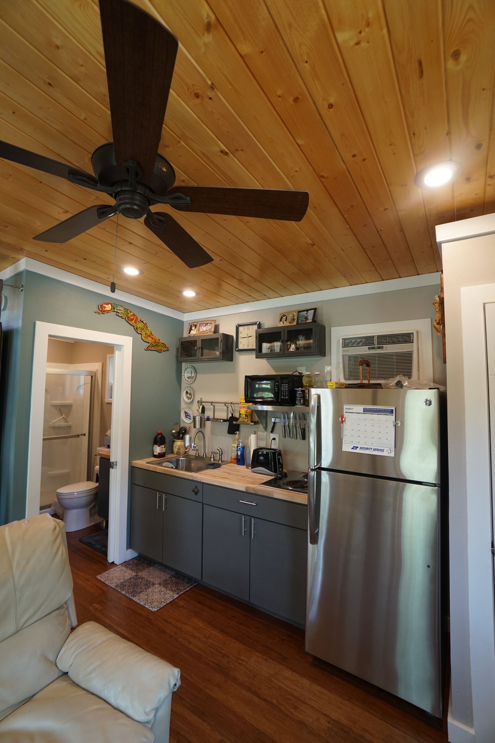 Small kitchen with stainless steel fridge, cabinets, microwave, and ceiling fan; bathroom entrance on the left.