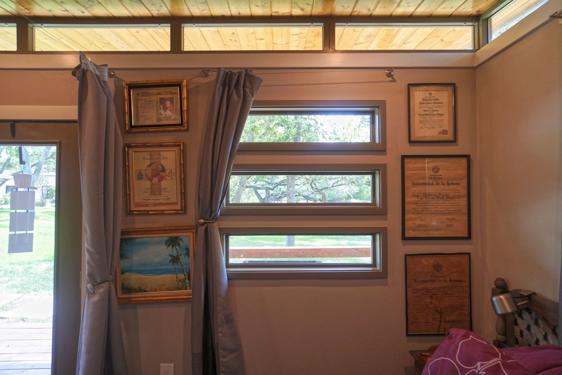 Interior view of a room with windows and framed artwork on a tan wall. Curtains hang beside a door.