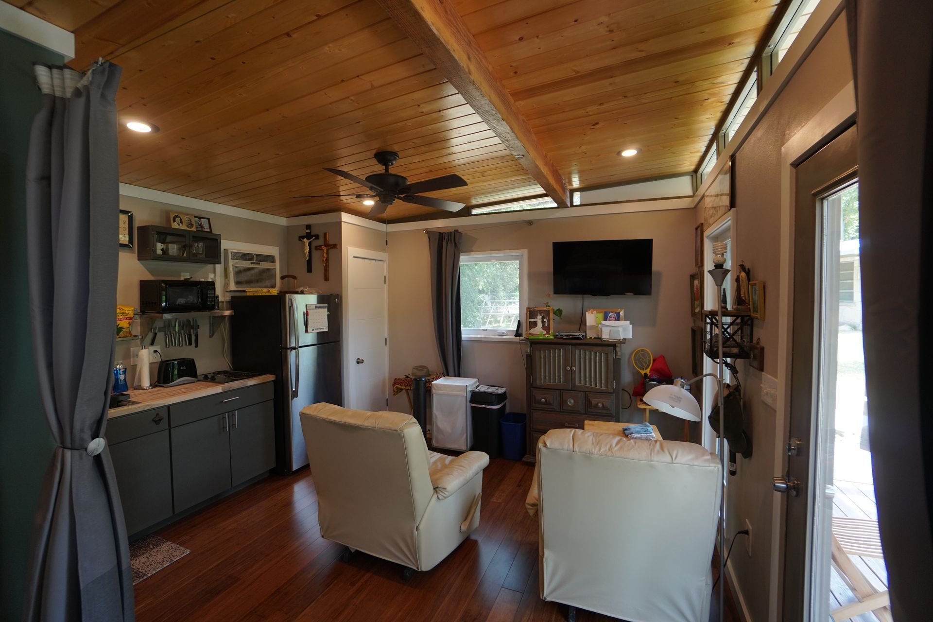 Interior of a cabin with wooden ceiling and flooring, featuring a kitchen, seating area, and television.