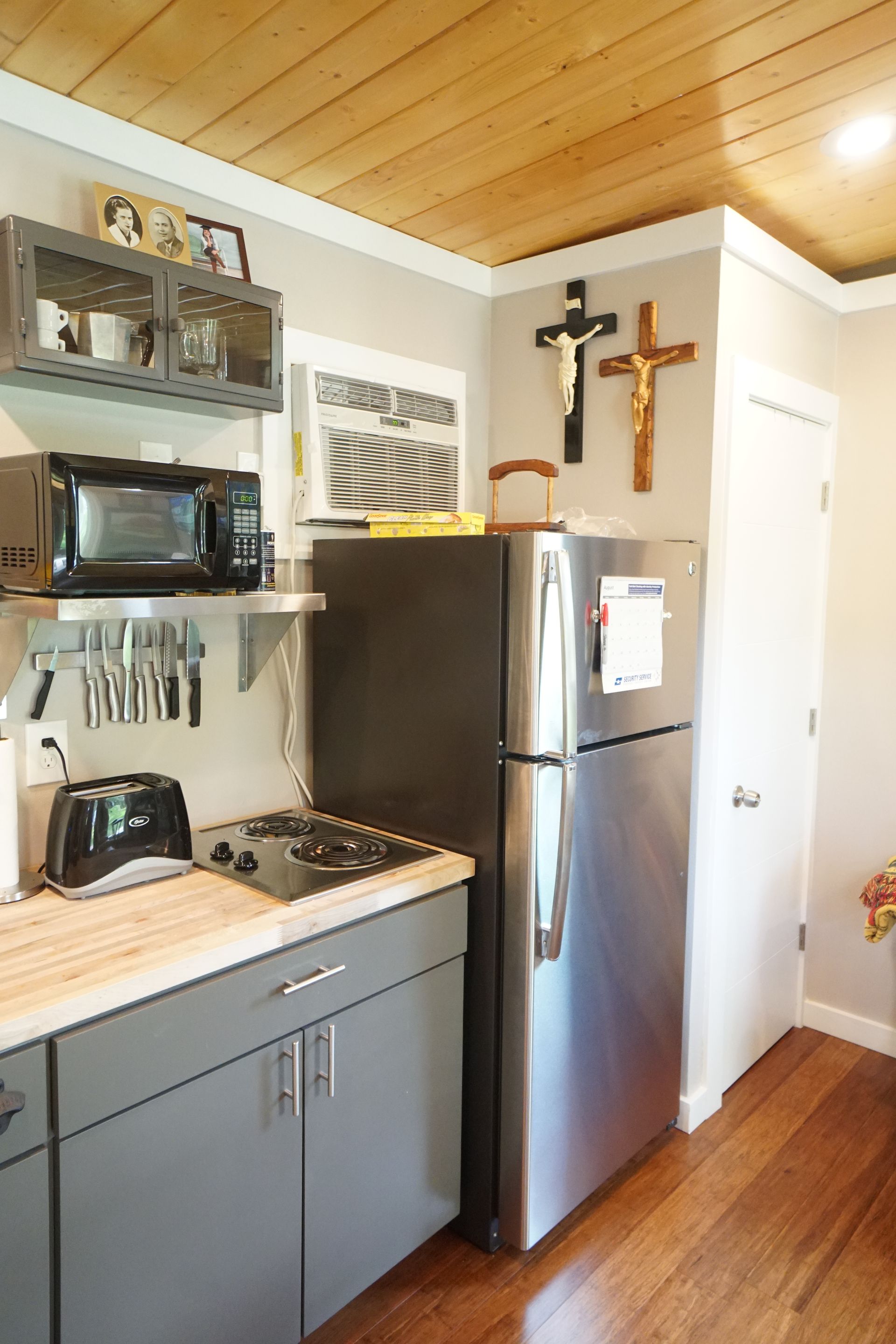 Small kitchen with grey cabinets, stainless steel appliances, and wooden ceiling. Two crucifixes hang on the wall.