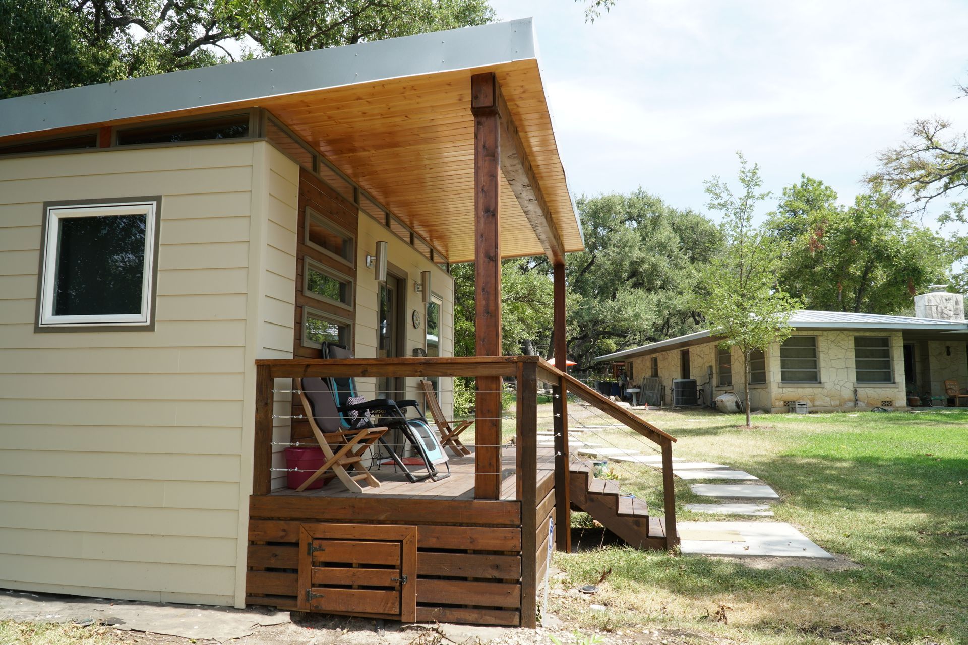 Tan and brown cabin with a porch and steps, set in a grassy yard, another building in the background.