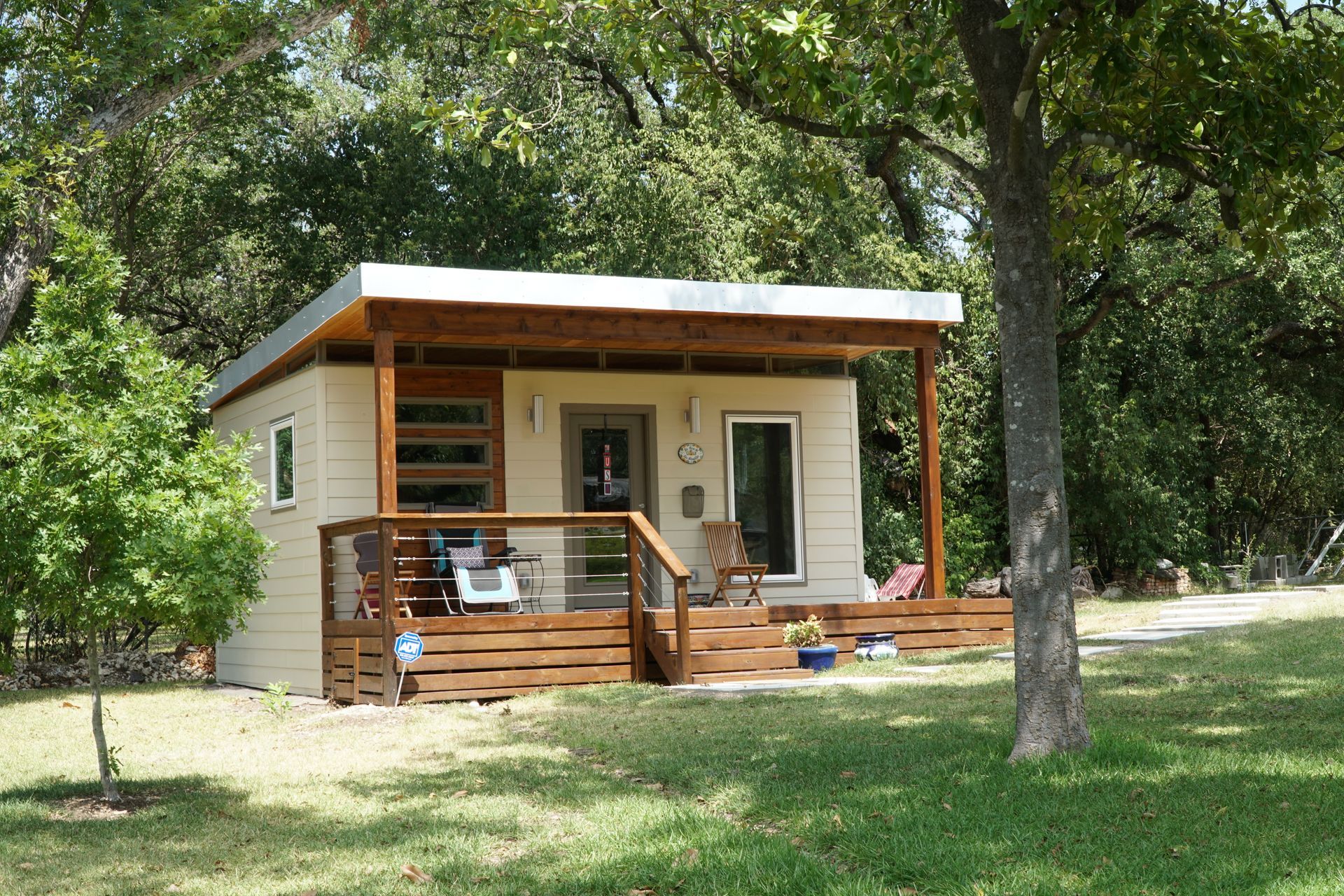 Small beige cabin with a wooden porch and a flat roof, surrounded by green trees on a grassy lawn.