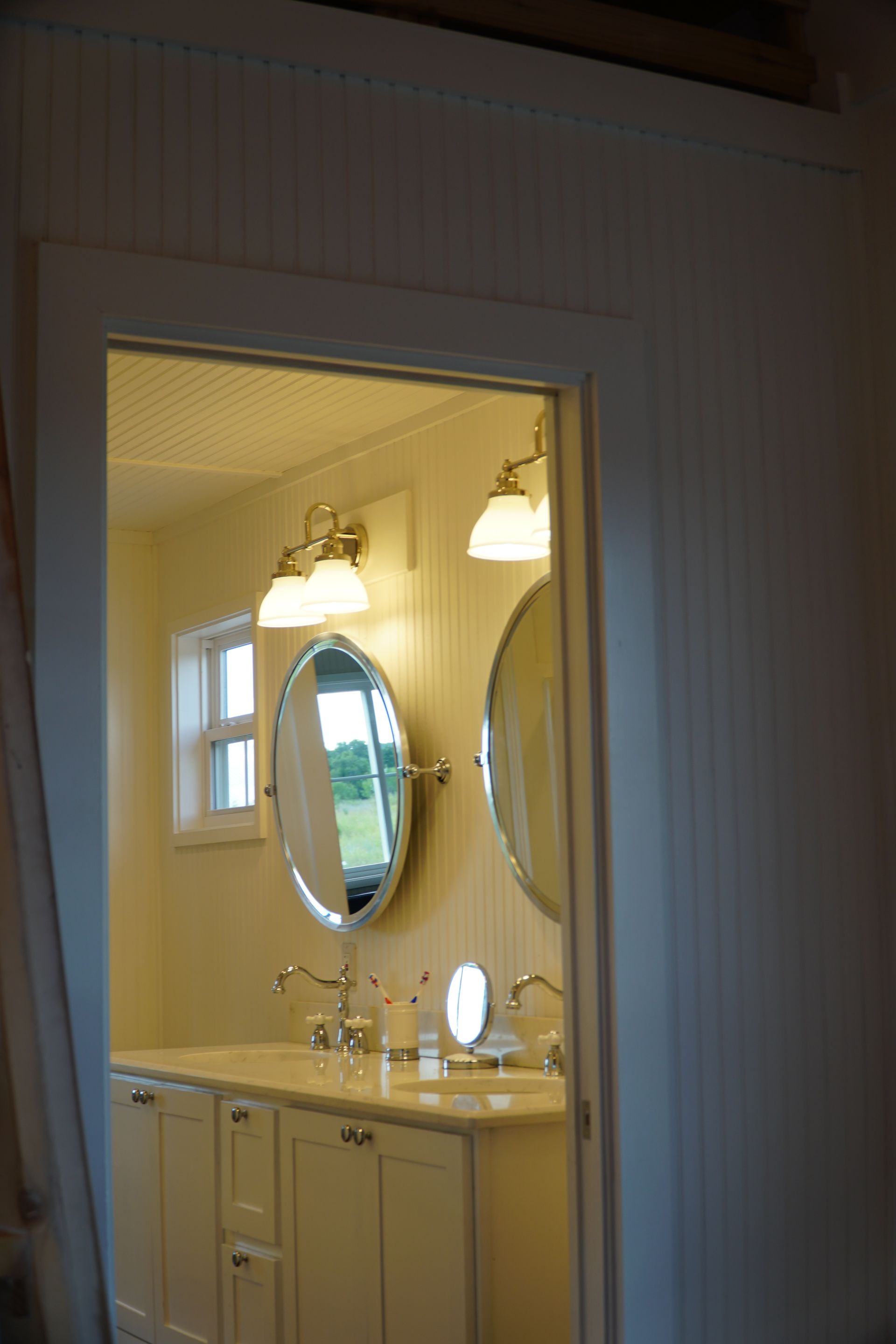 Bathroom interior, viewed through a doorway. Oval mirrors, vanity with sinks, and sconces are visible.