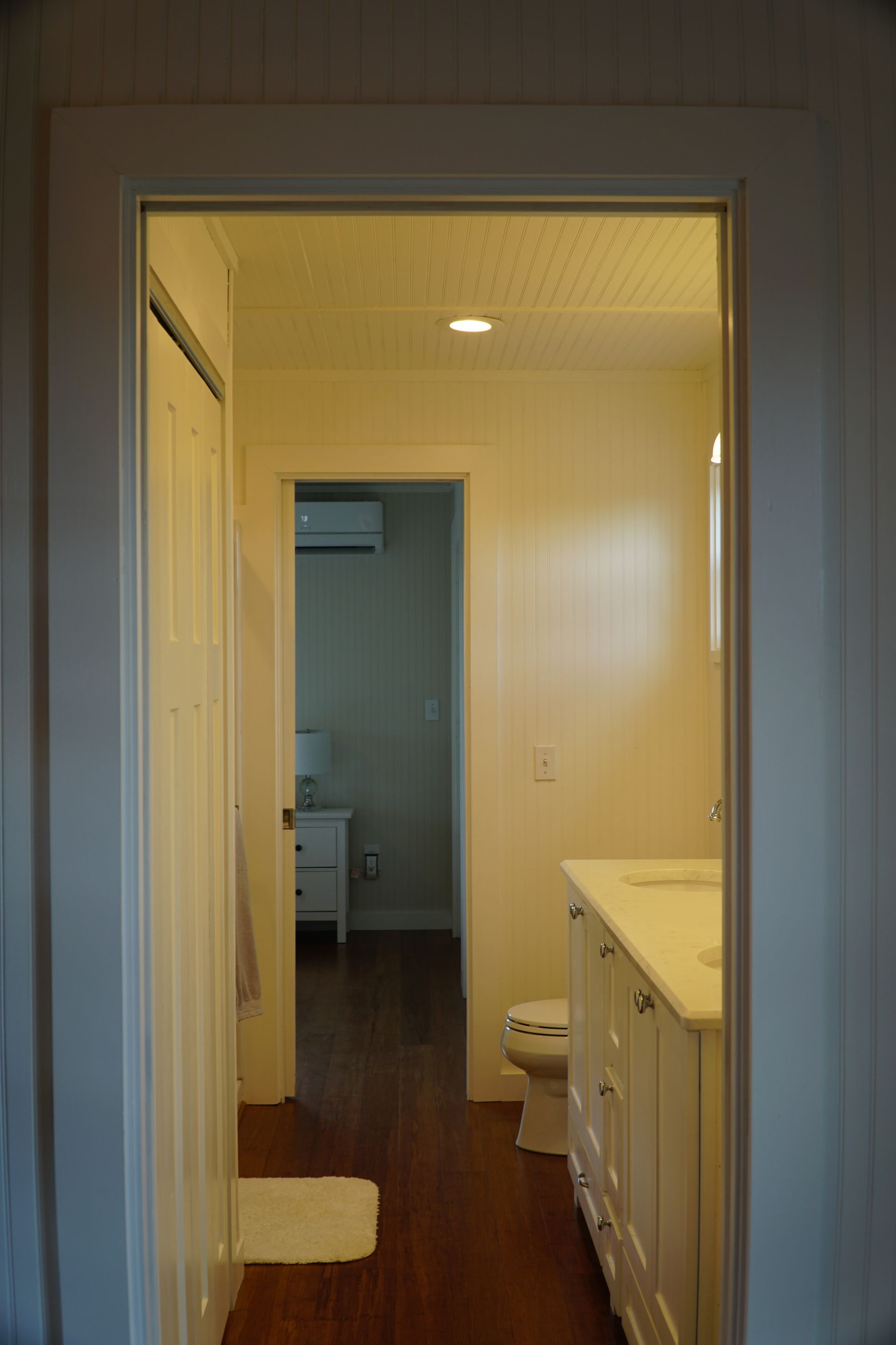 View through a doorway into a bathroom and a bedroom; white walls and trim, wood floor.