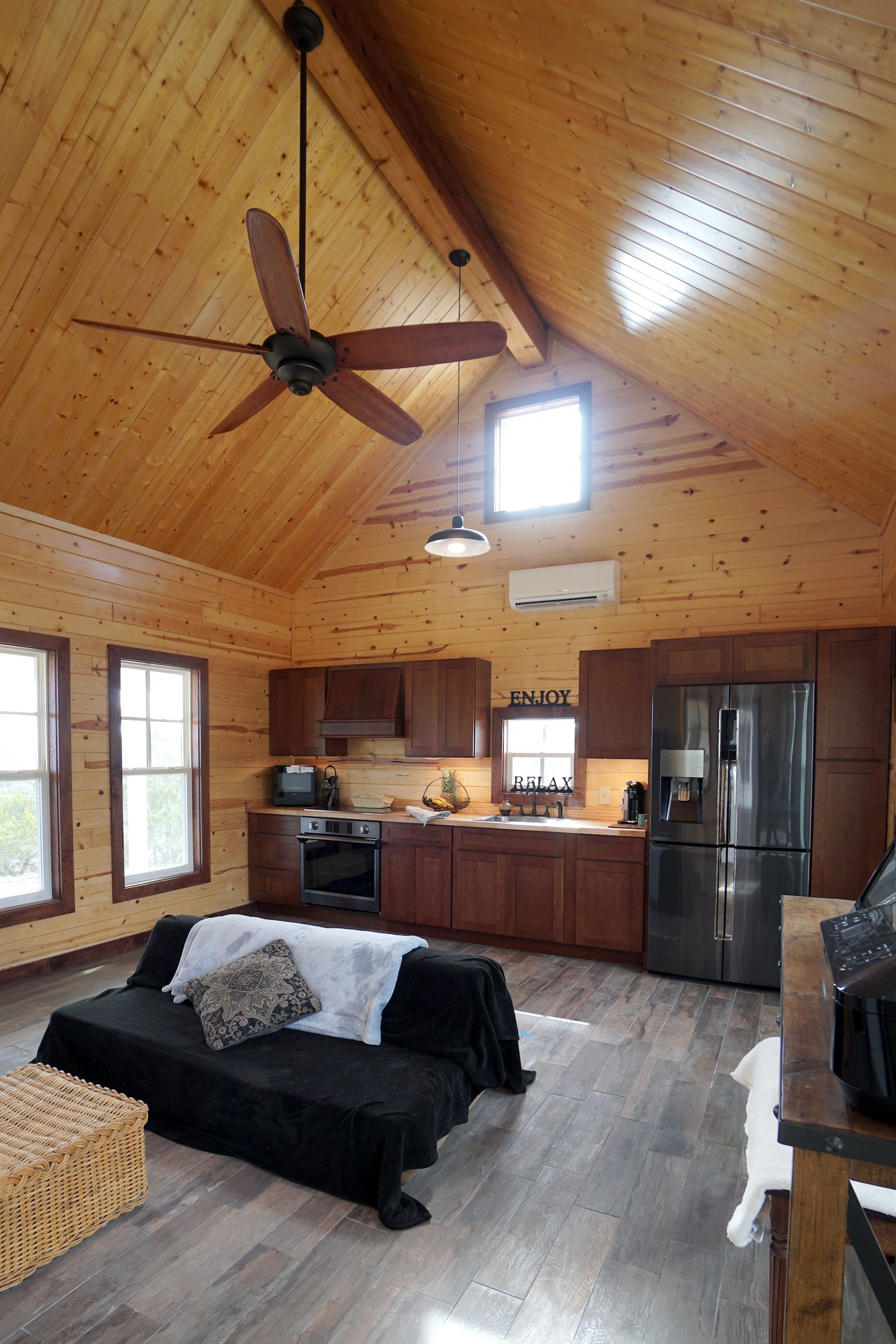 Interior of a cabin with wood walls and ceiling. Kitchen with dark cabinets, sofa, and a ceiling fan.