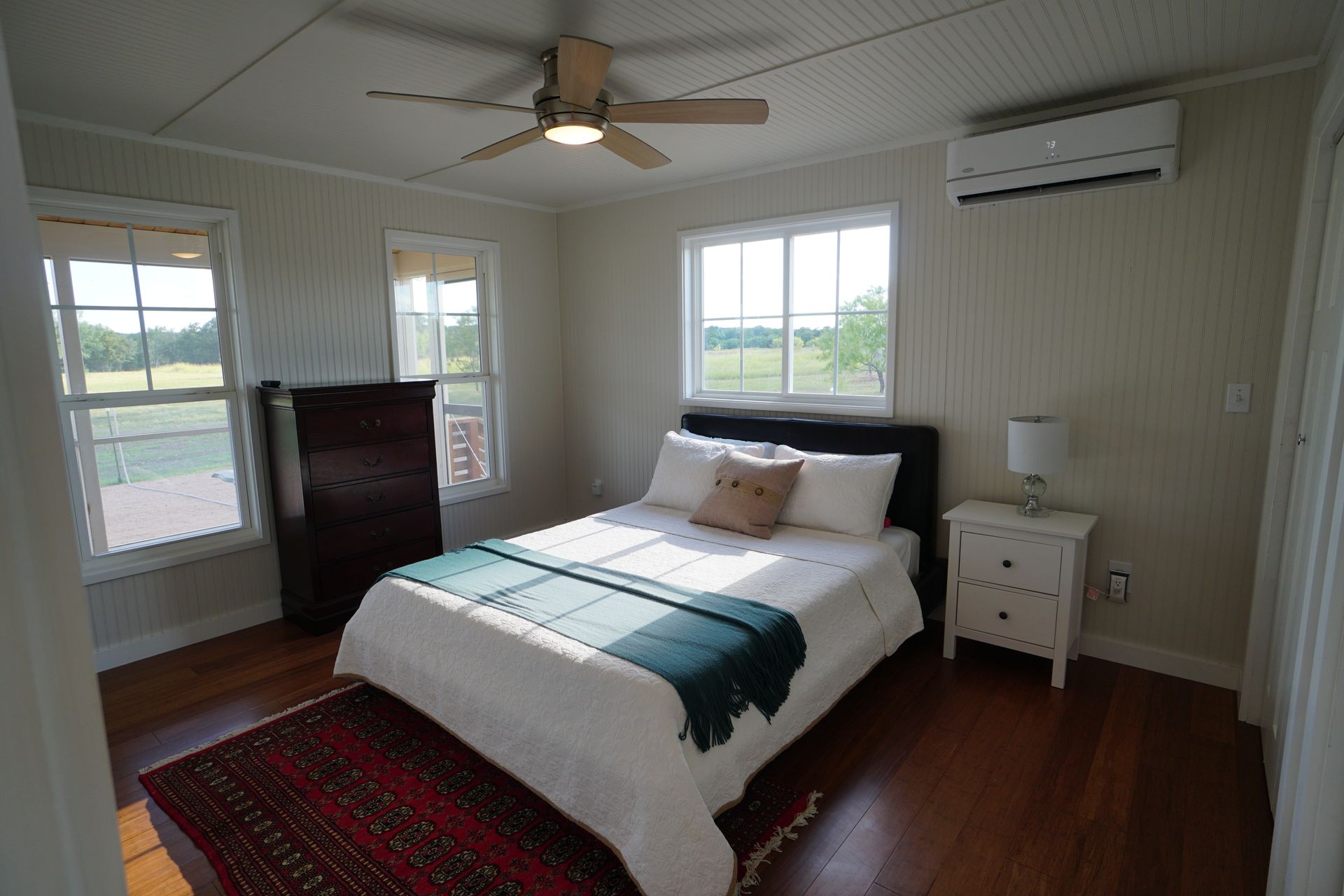 Bedroom with a bed, dresser, nightstand, windows, and ceiling fan. Wooden floor and white walls.