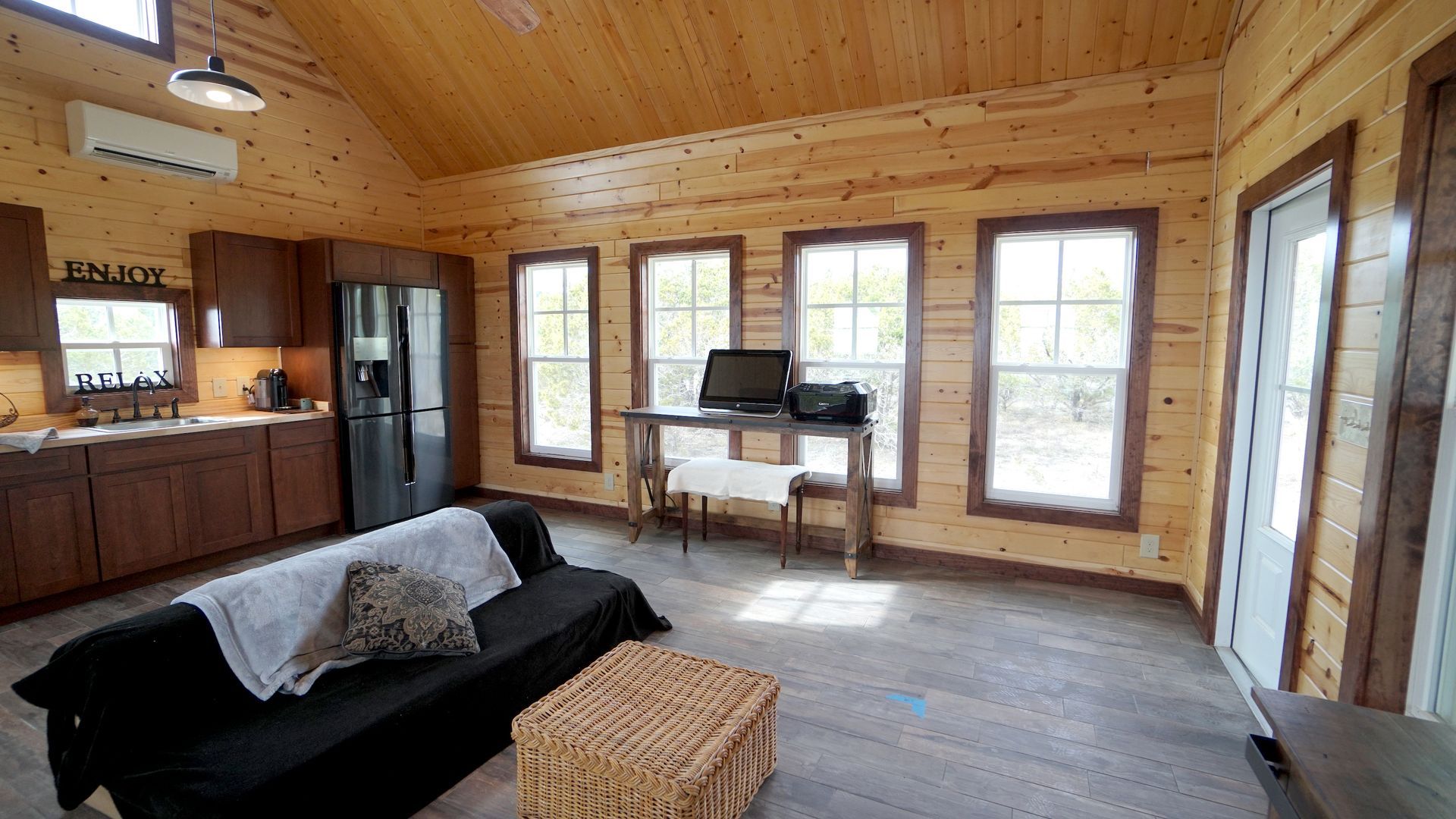 Cozy cabin interior with wood-paneled walls and ceiling.  Living area with sofa, kitchen, desk by the windows.