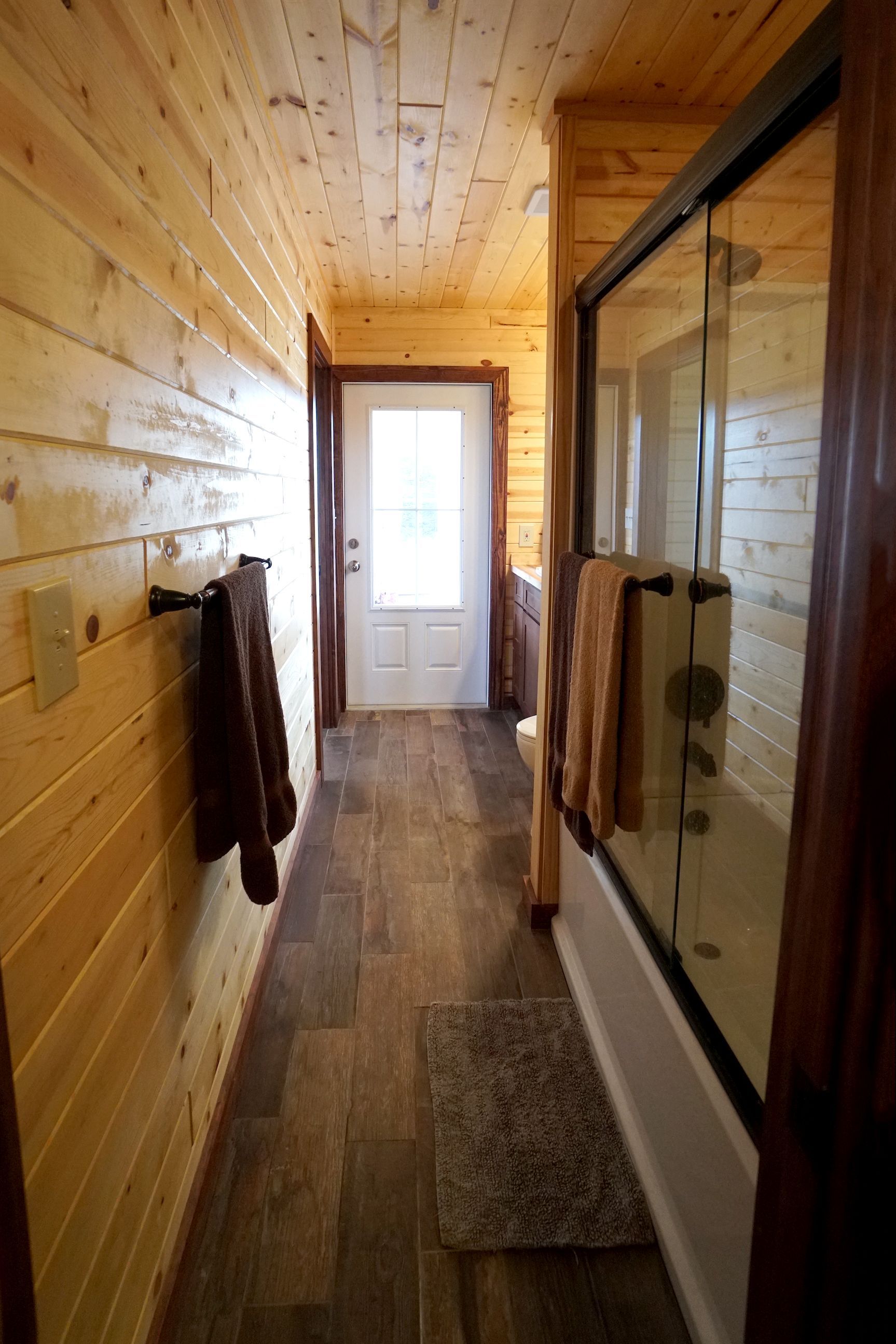 Narrow bathroom with wood paneling, door, shower, and towels. Brown and beige tones.