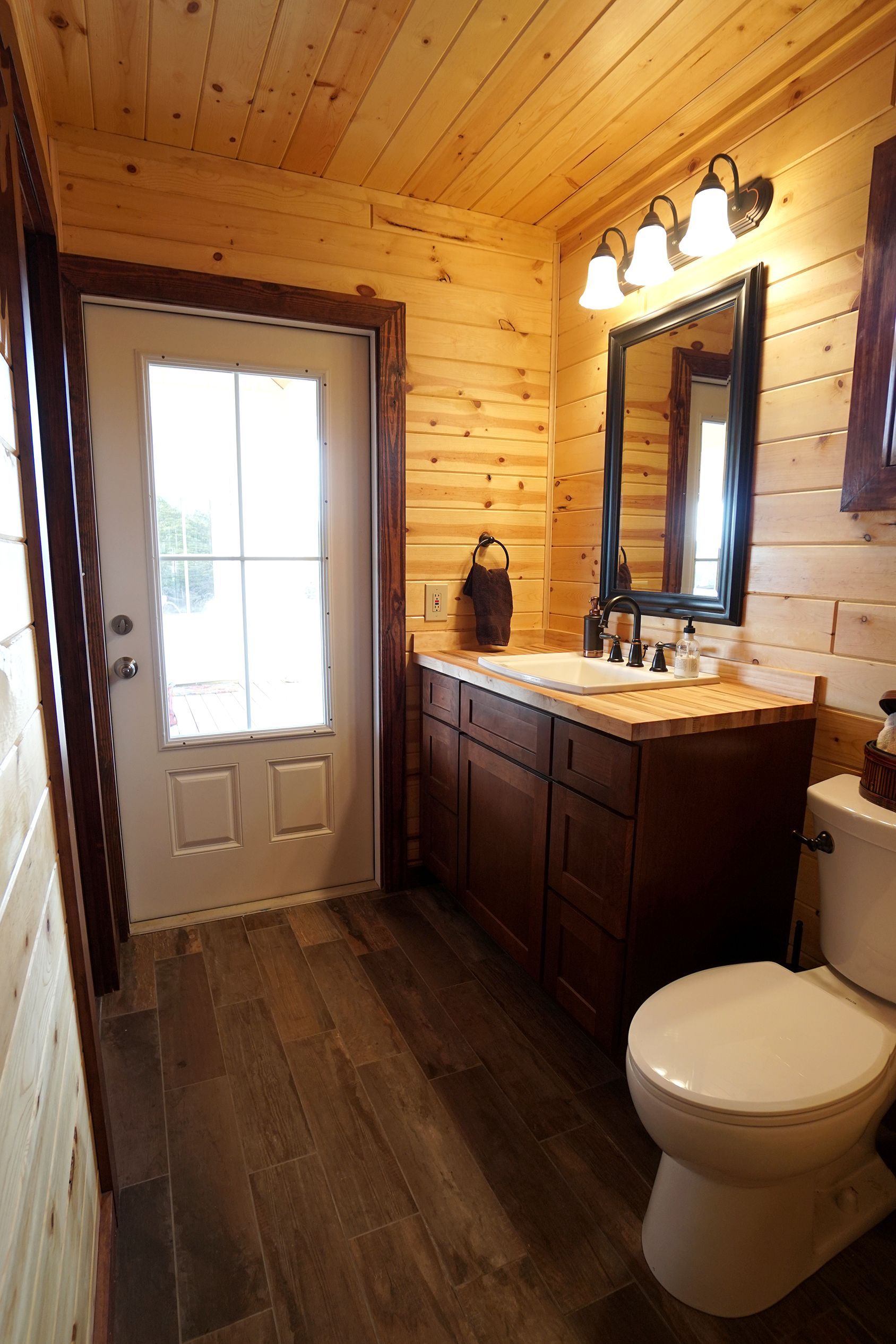 Bathroom with wood-paneled walls, a vanity with a mirror, a toilet, and a door leading outside.