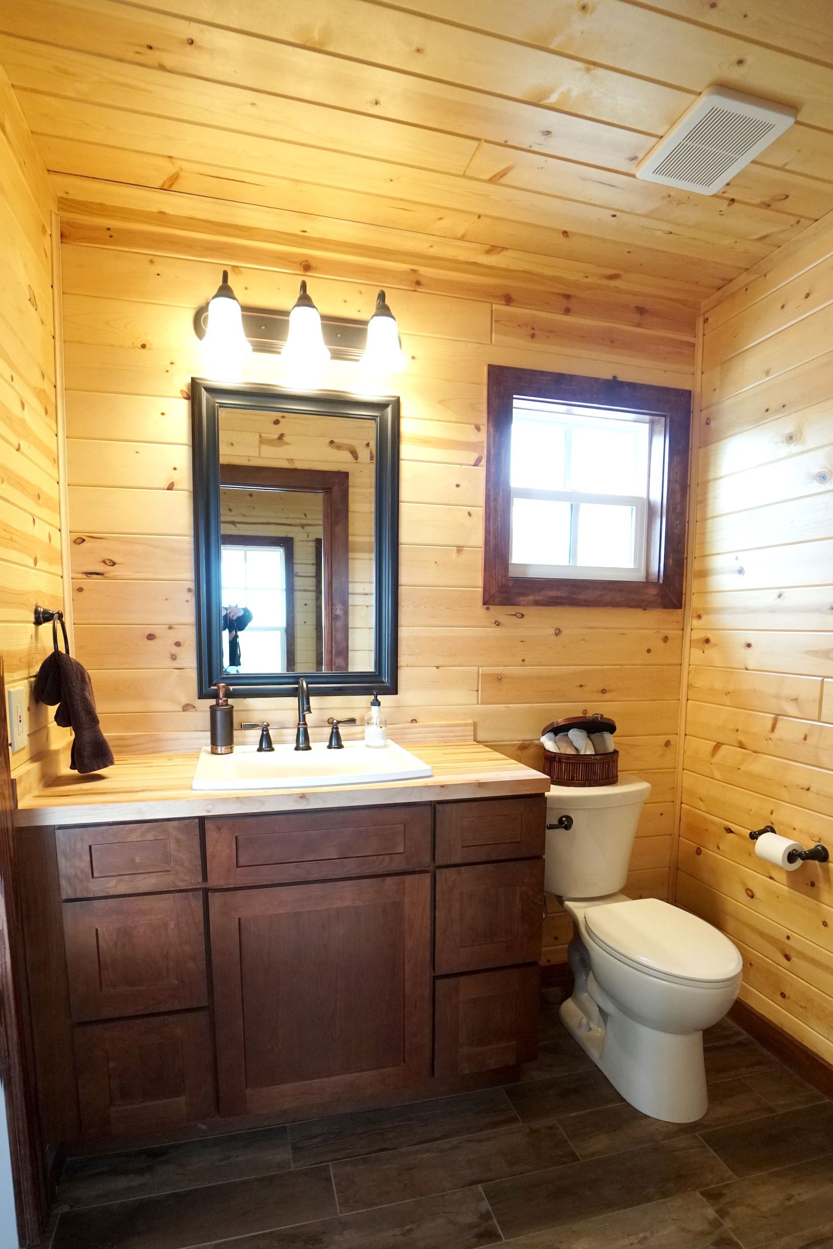 Bathroom with wooden walls and ceiling, brown vanity, toilet, mirror, and a small window.