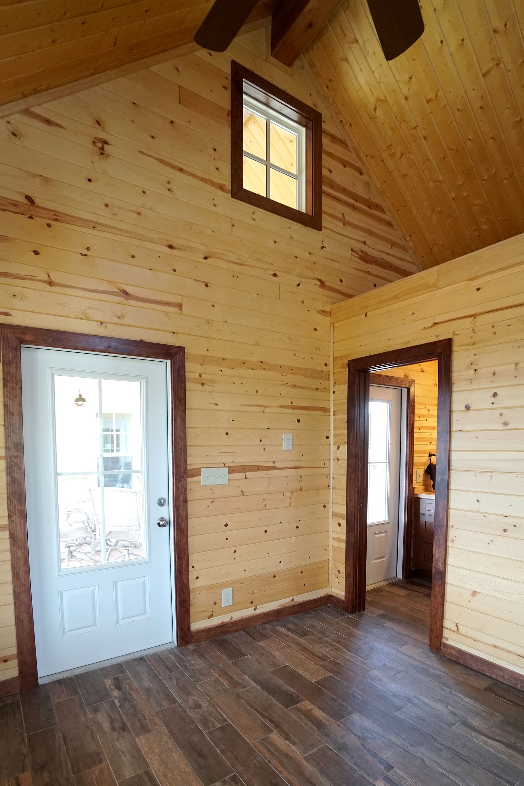 Interior of a small cabin with wooden walls, dark floor, white door, and a small window.