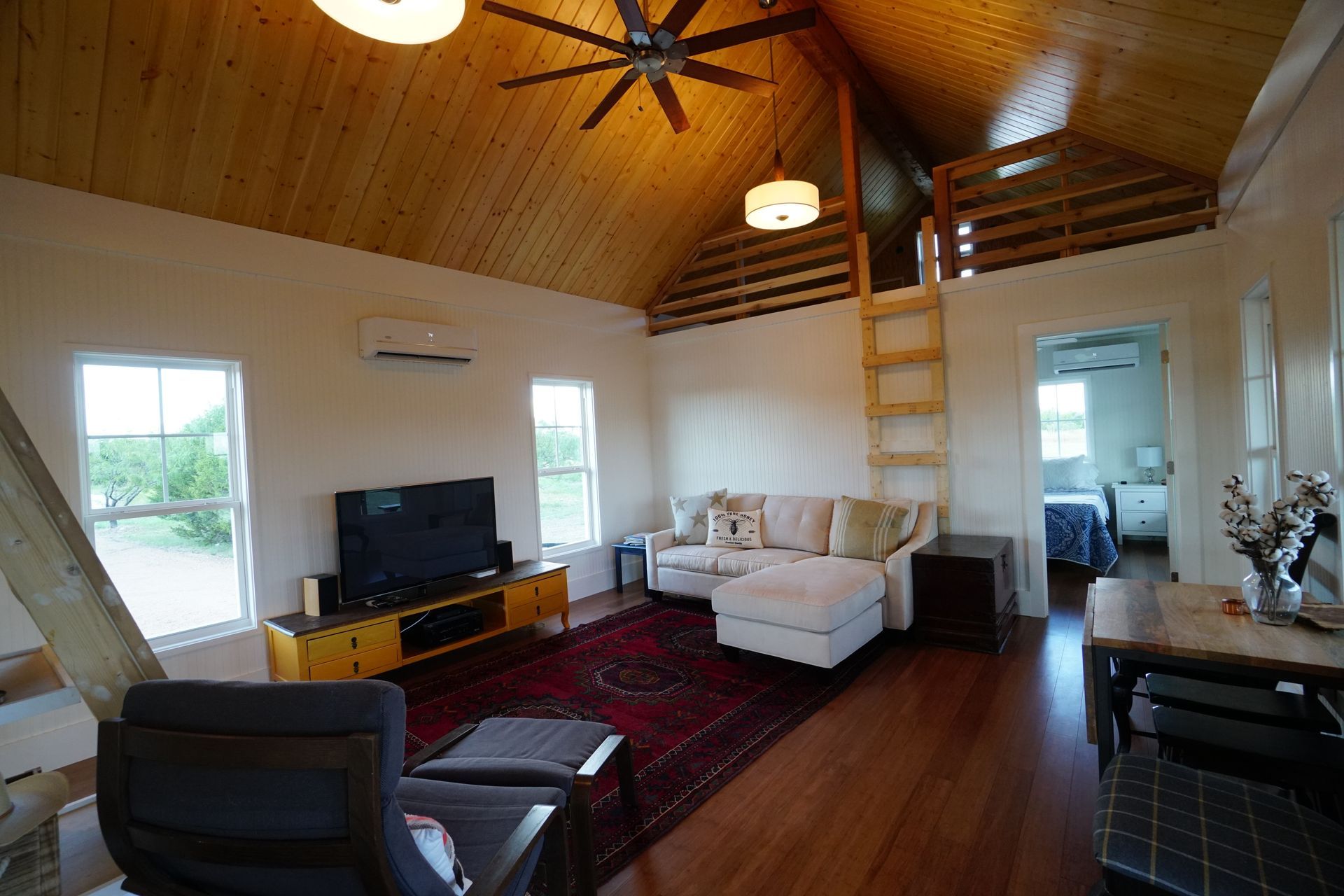 Living room with wooden ceiling, sofa, TV, rug, and loft accessed by a ladder.