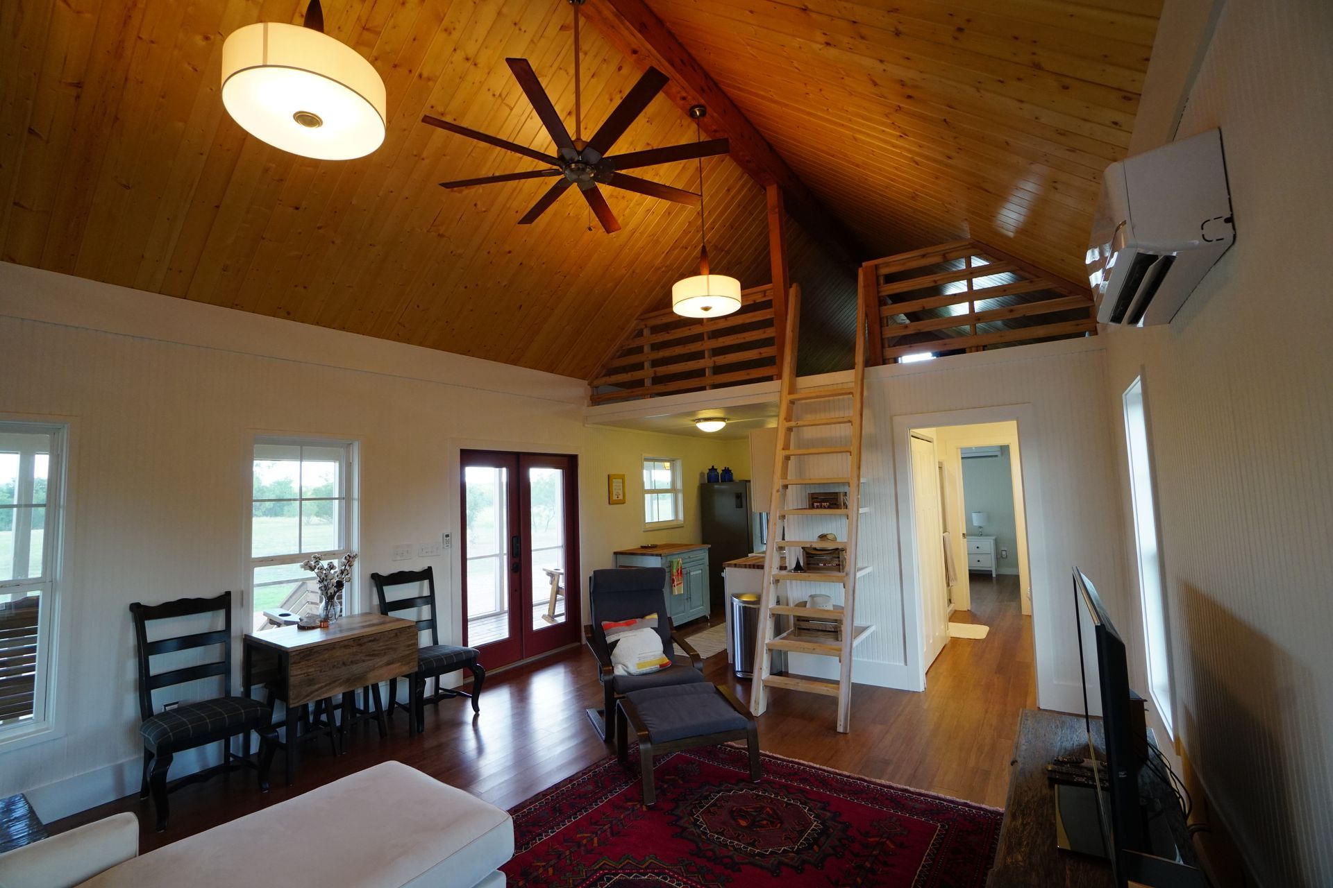 Bright, open living space with wood ceiling, loft, and red rug. Includes chairs, table, and a ladder.