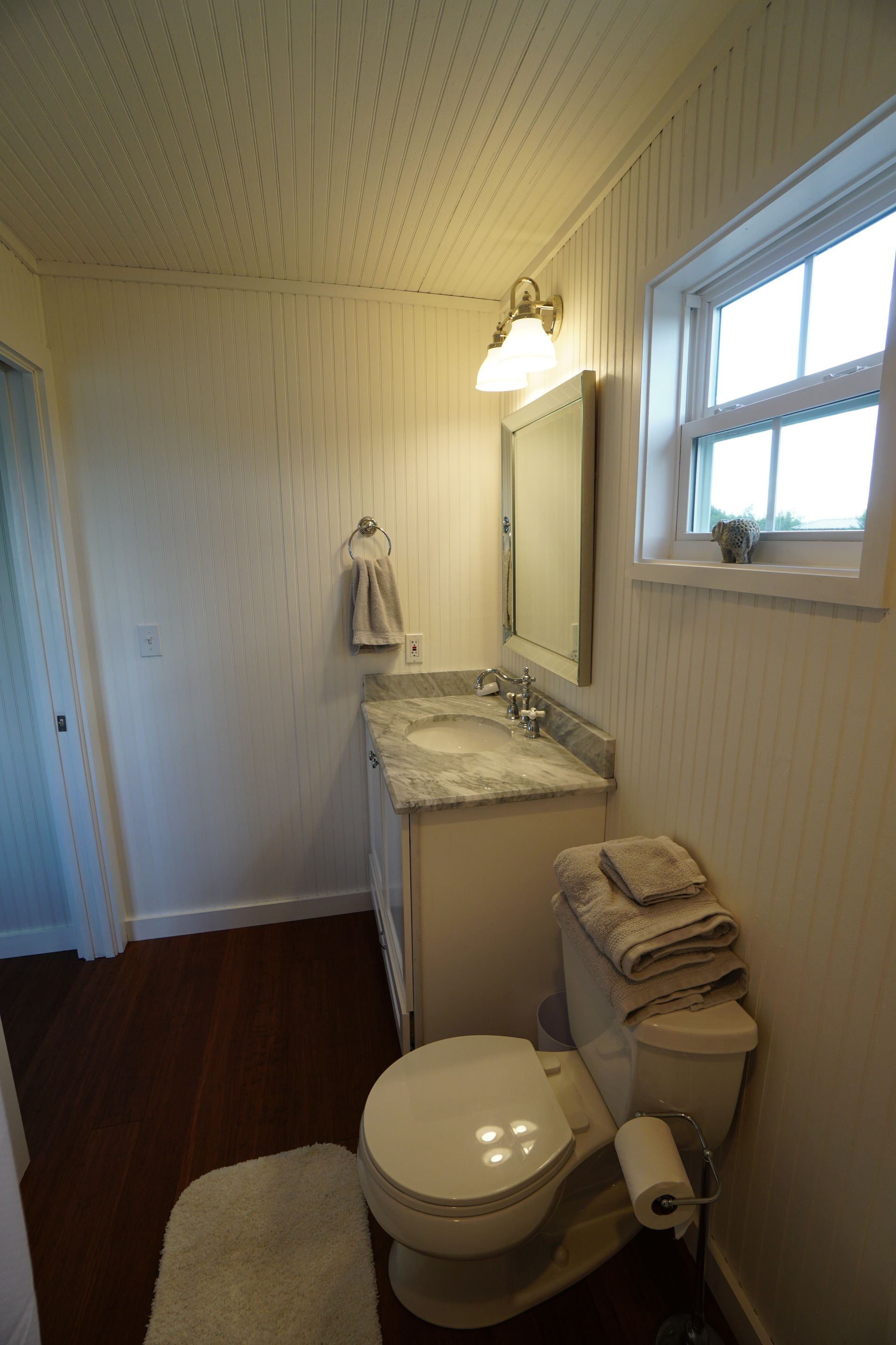 Bathroom with white walls, vanity, toilet, and window. Brown floor.