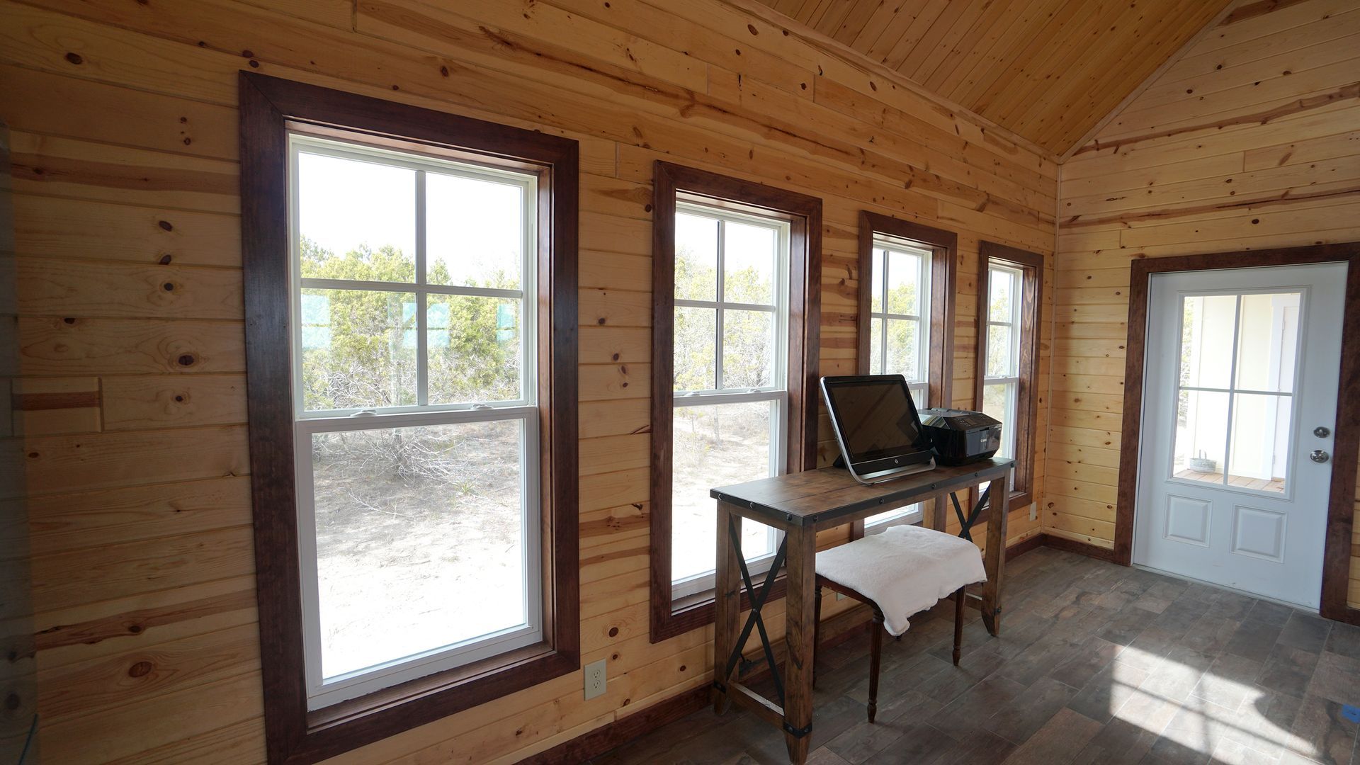 Interior of a small room with wood walls, three windows, a desk with computer, and a door.