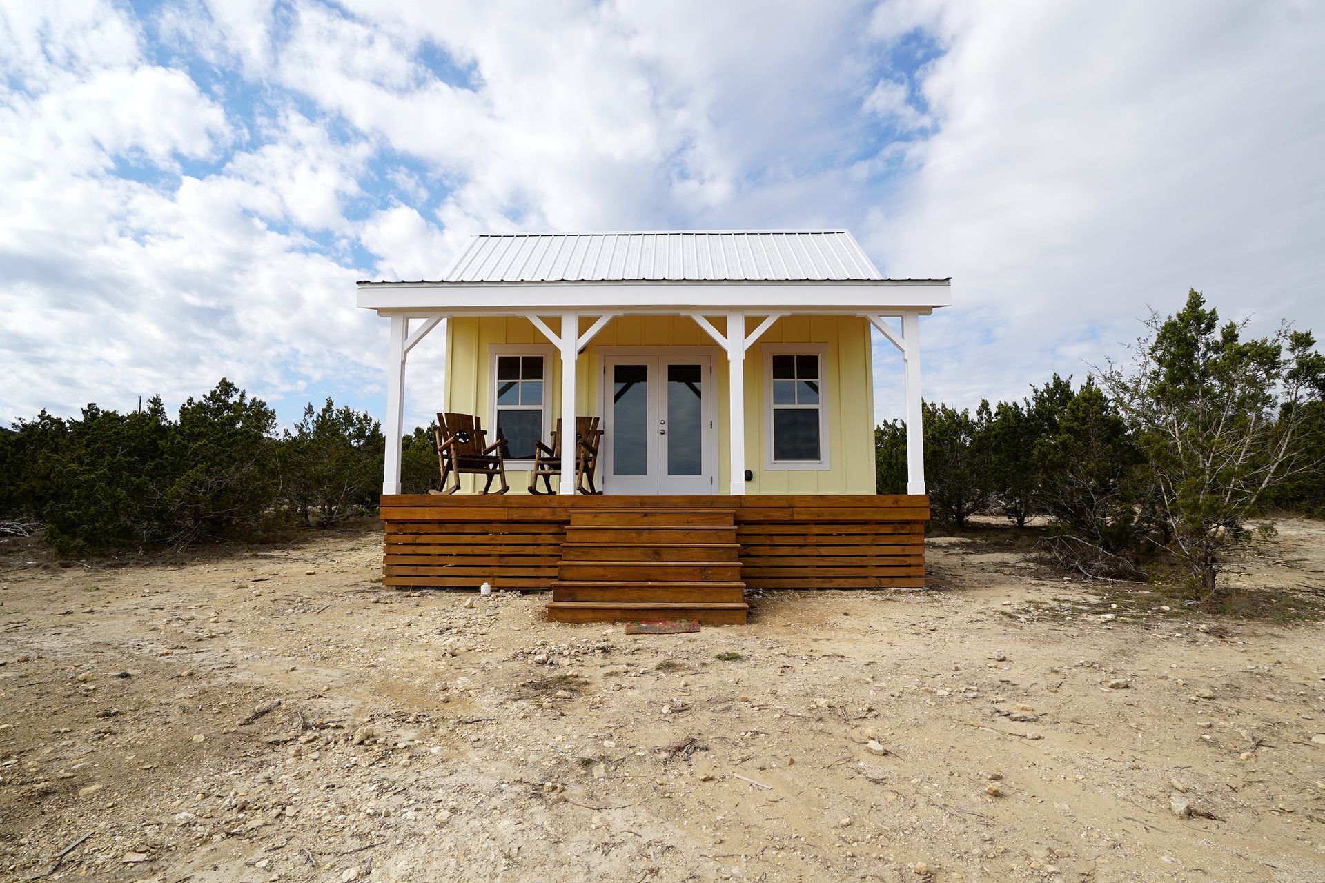 Yellow tiny house with white porch and stairs on a sandy lot under a cloudy sky.