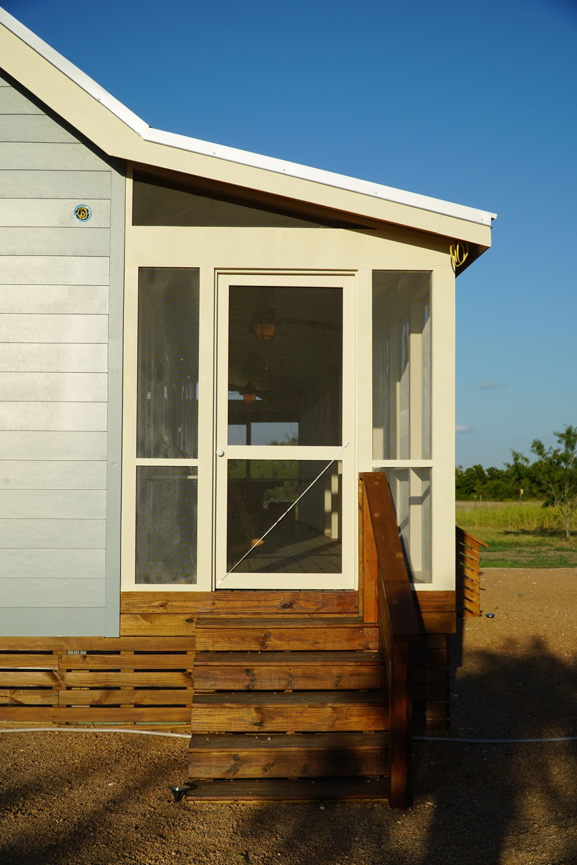 Wooden steps lead to a screened-in porch with a door; light blue siding on the left, clear blue sky.