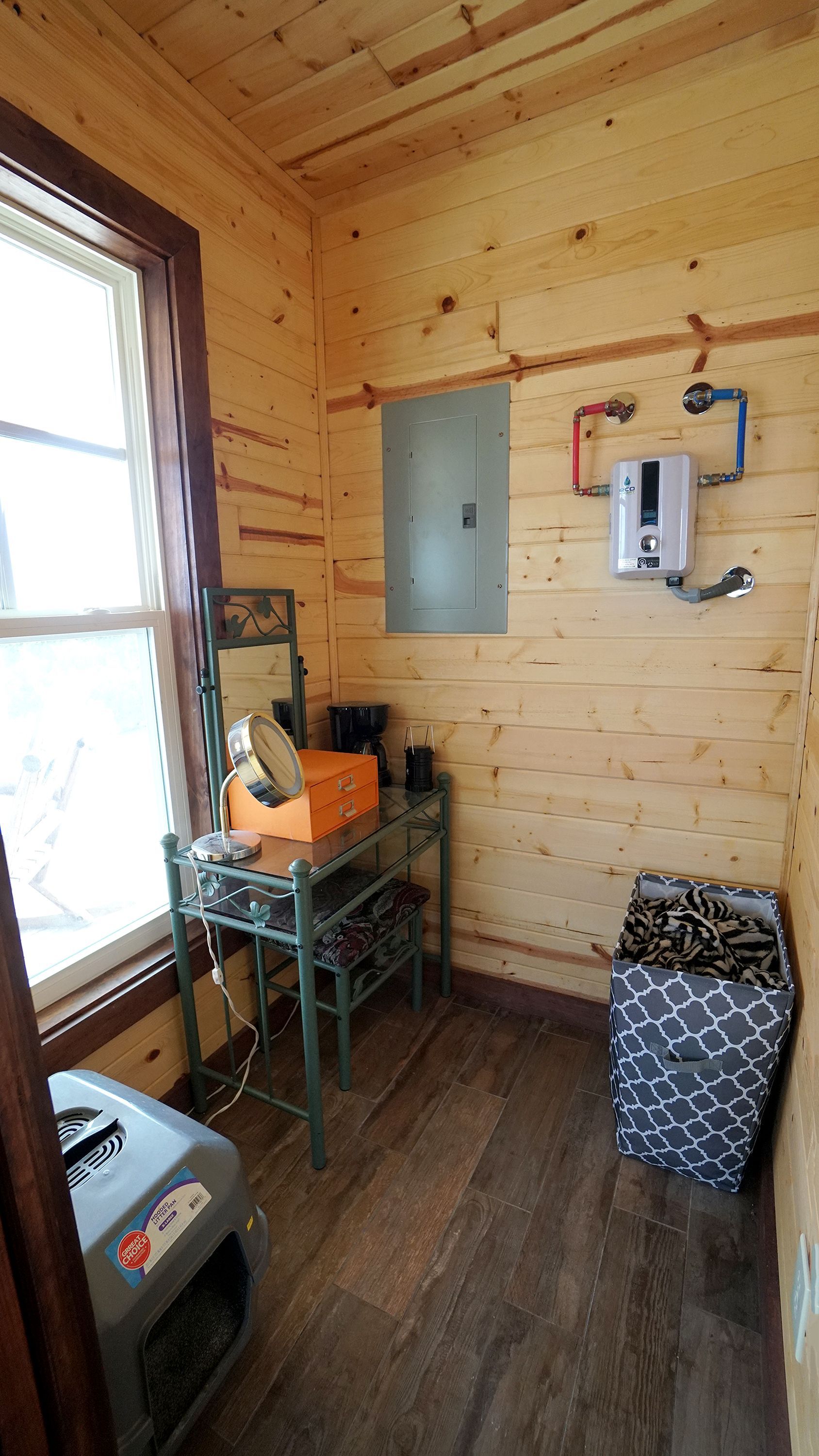 Small room with wood walls and floor, window, electrical panel, water heater, and storage bin.