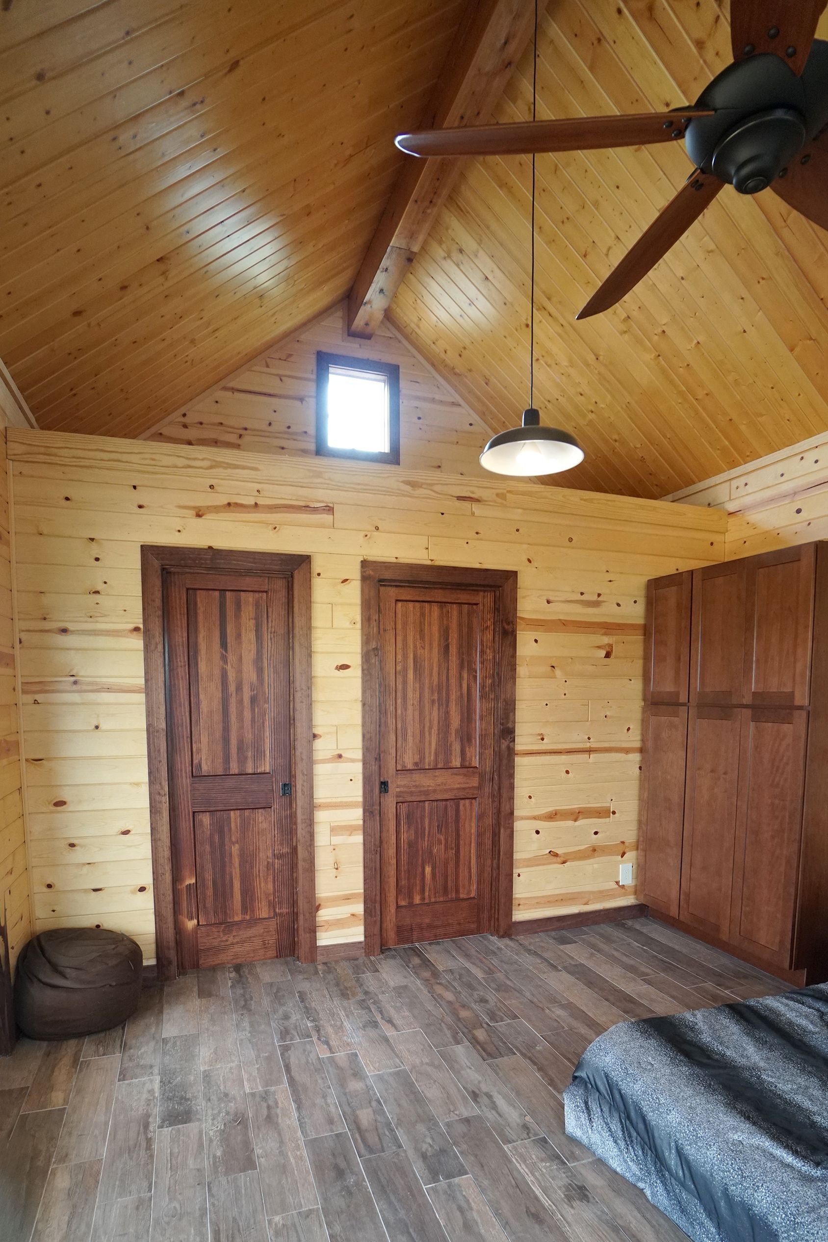Interior of a wooden cabin bedroom: wooden walls, doors, ceiling fan, and a small window.
