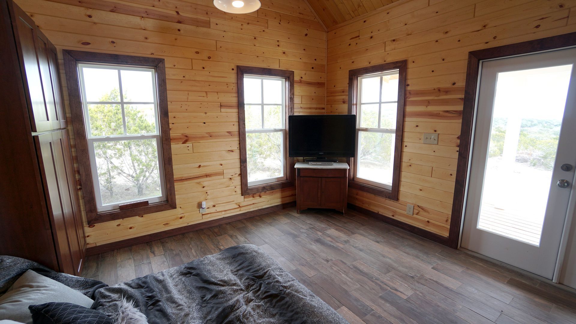 Wooden-walled room with windows, TV, and door. A bed is in the foreground.