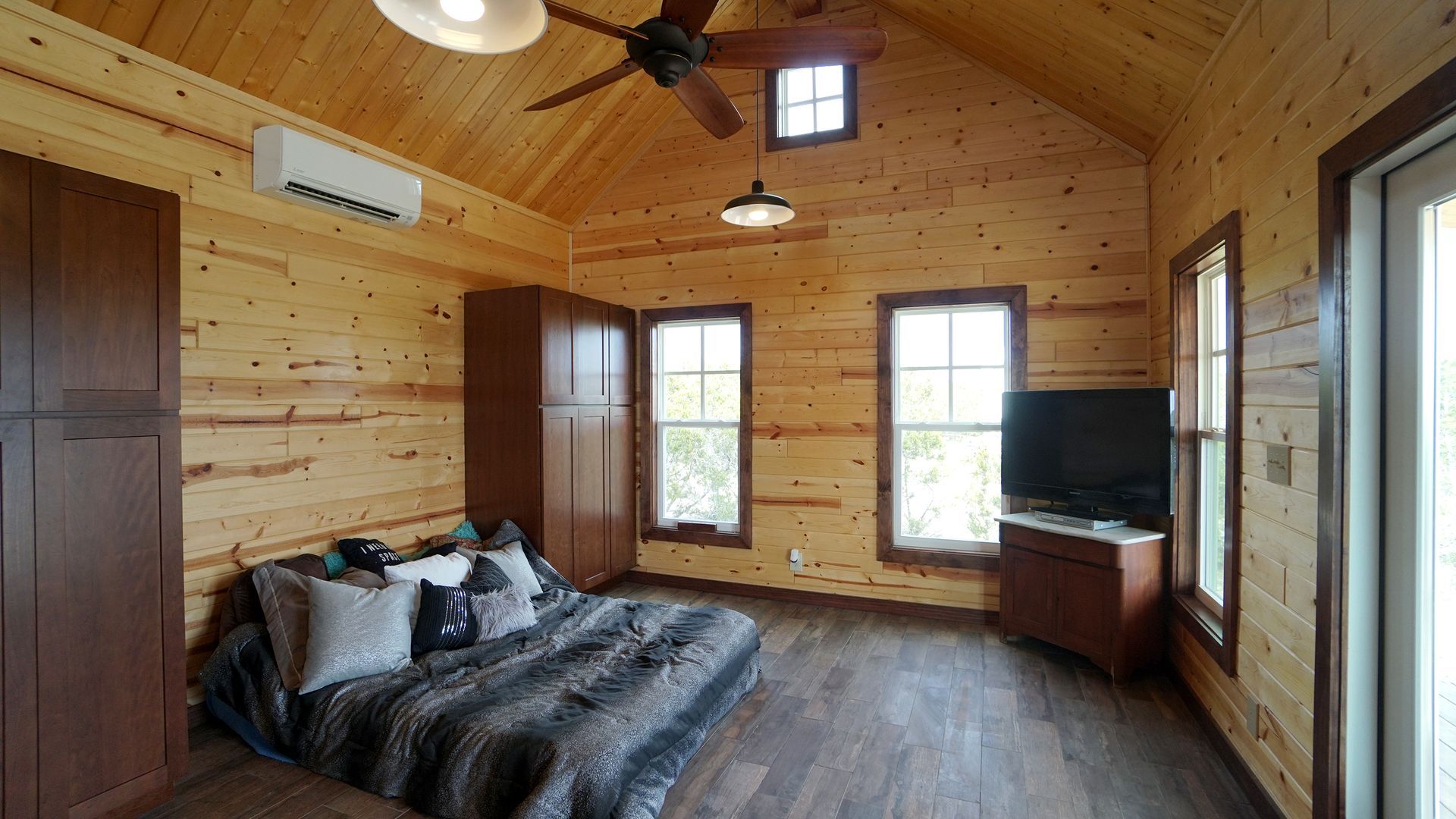 Cozy cabin interior with wood paneling, couch, TV, and ceiling fan. Natural light streams from the windows.