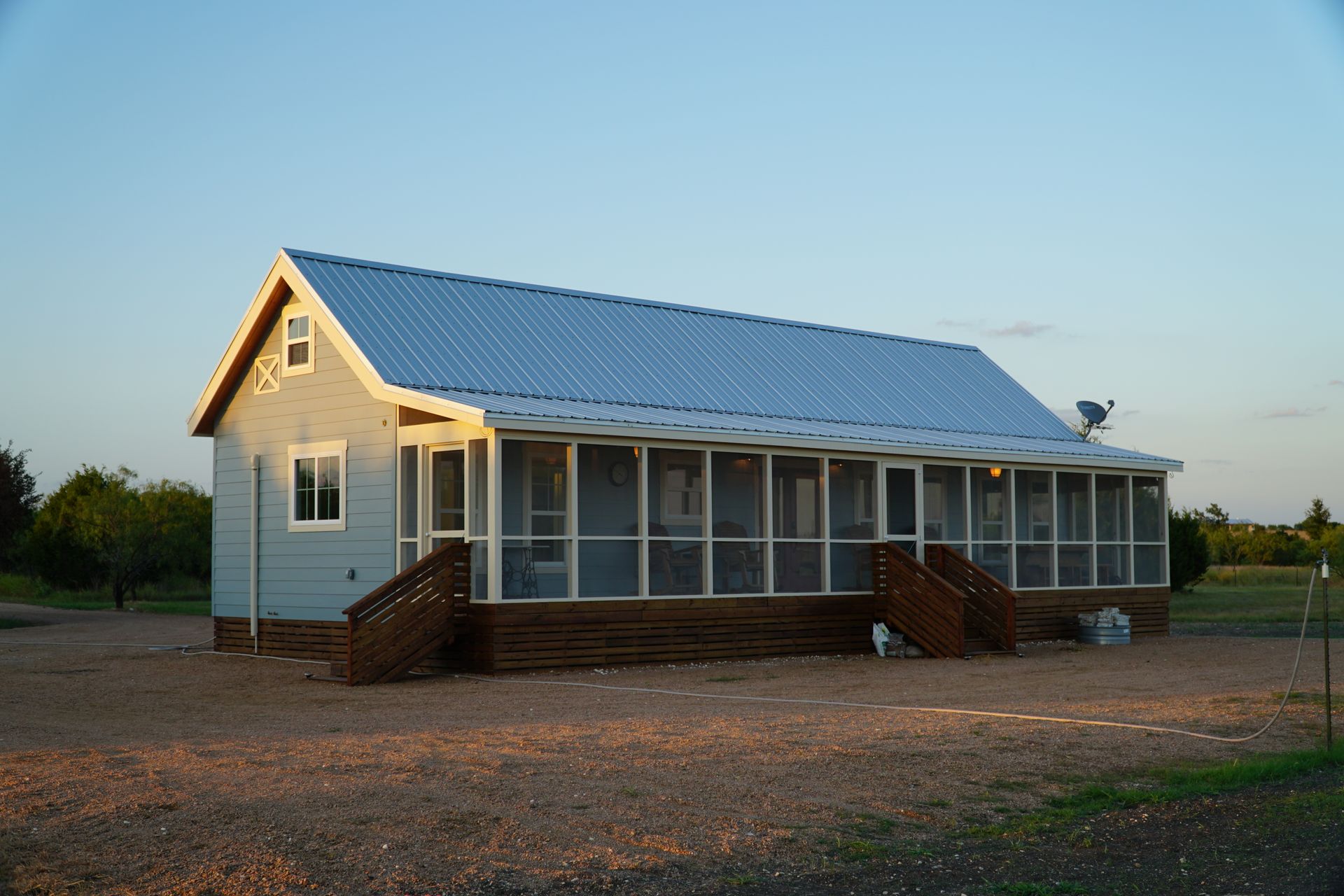 Light blue cabin with screened-in porch, metal roof, wooden steps, set in a gravel yard, under a blue sky.