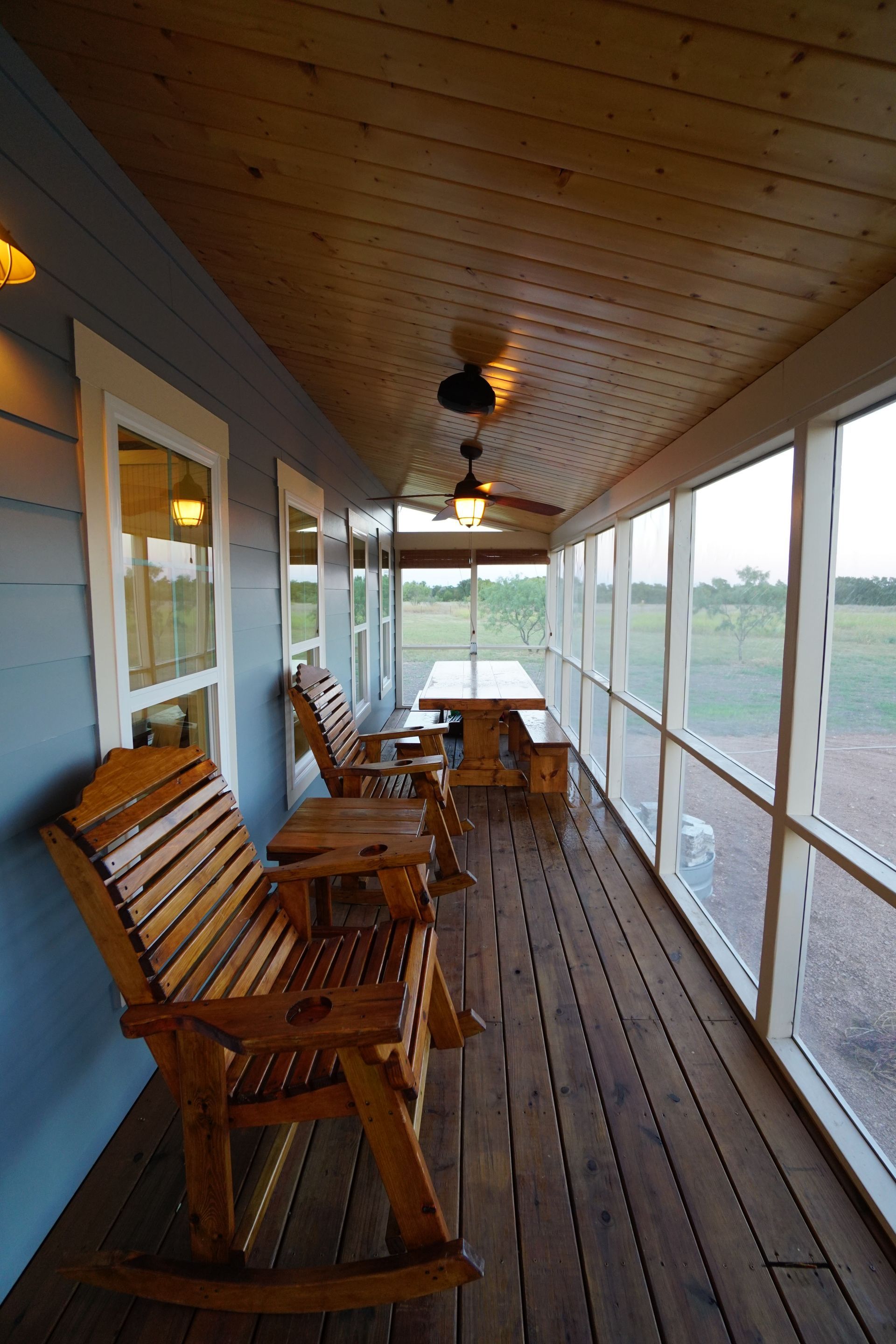 Screened porch with wooden rocking chairs and a table, overlooking a grassy field.